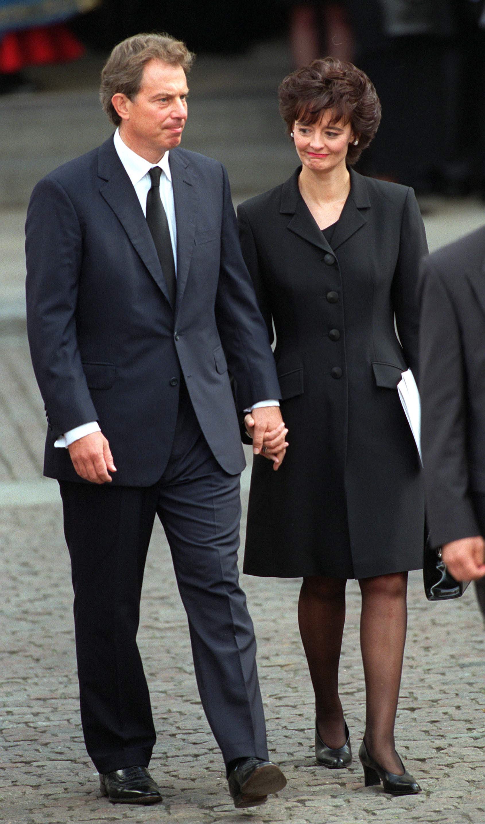 Two people walk hand in hand, dressed in formal black attire, on a cobblestone street with others blurred in the background.