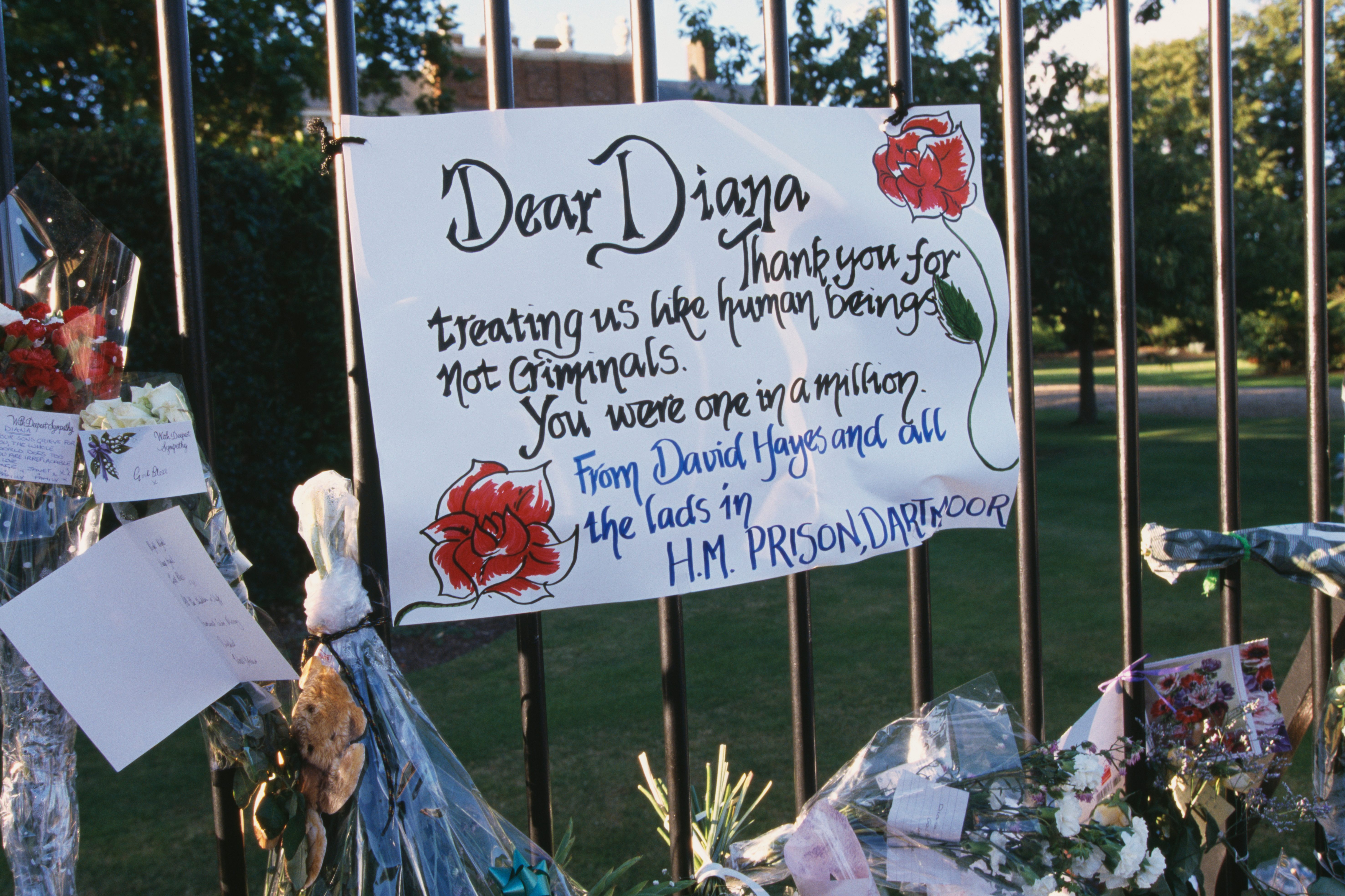 A handwritten note thanking Diana is displayed on a black fence, surrounded by flowers and cards, in a park-like setting.