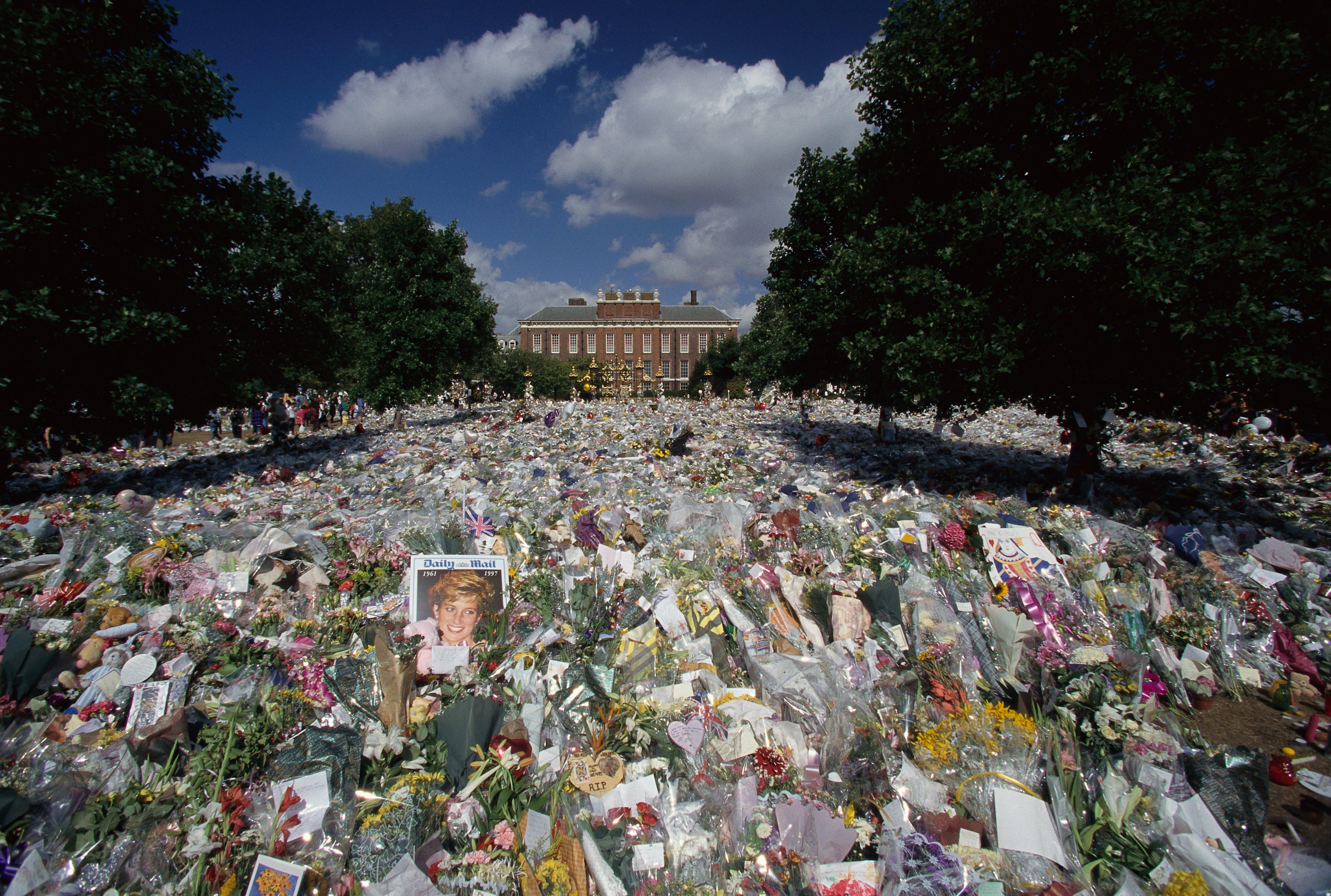 Flowers cover the ground in tribute outside a large building, surrounded by trees and blue sky, with notes and photographs visible.