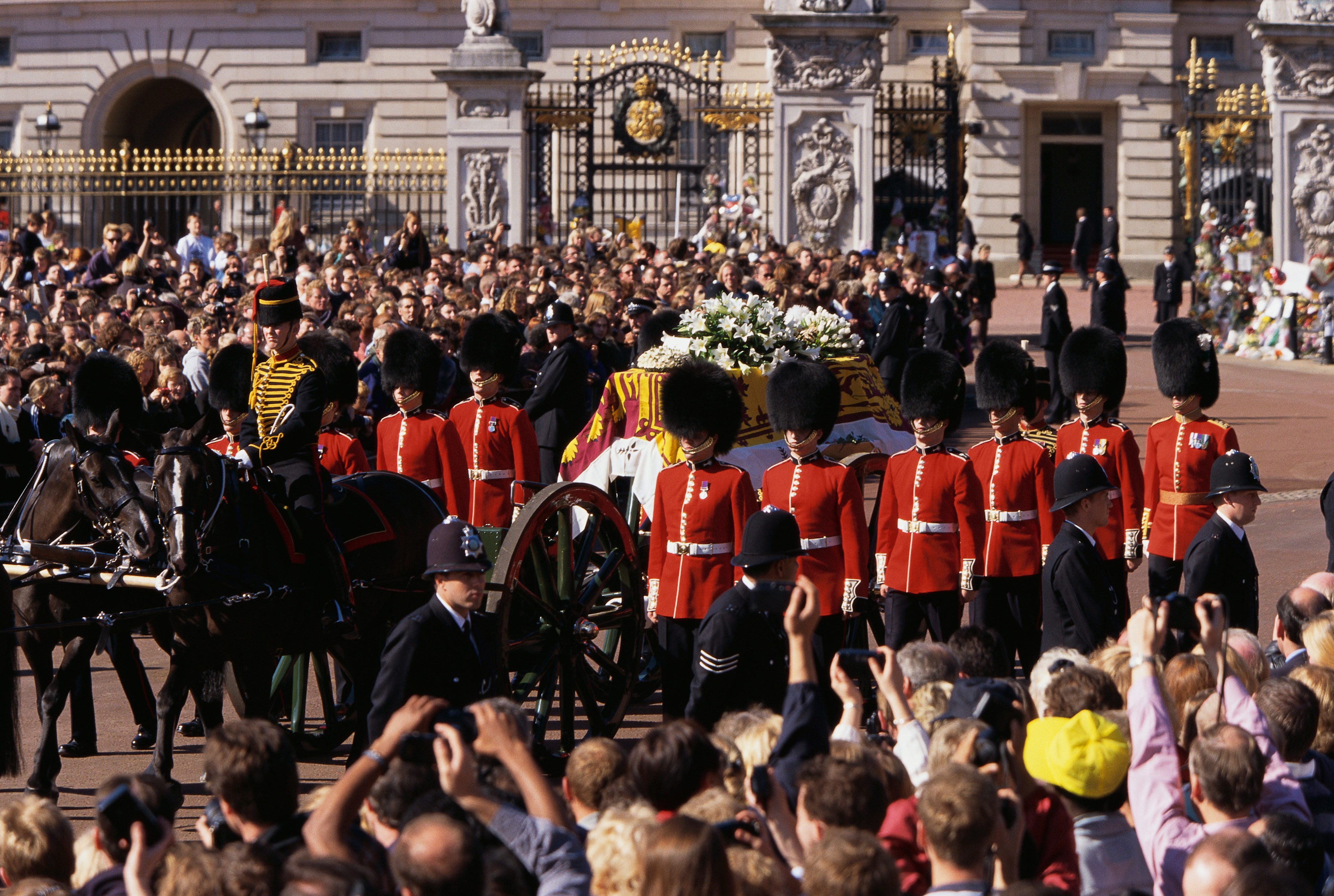 Casket draped in a floral-covered flag moves in a procession, escorted by uniformed guards, before a large crowd outside a historic building.
