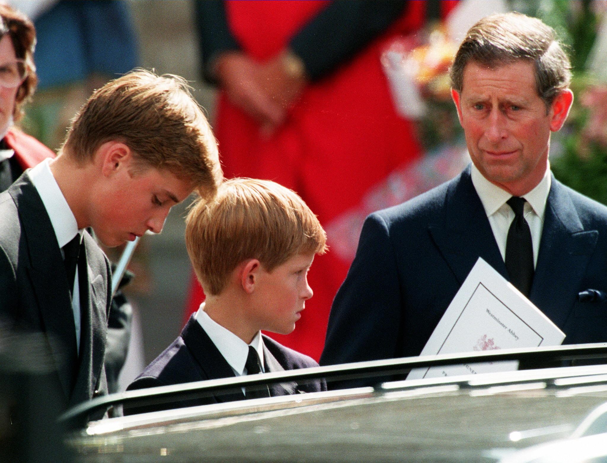 Three people dressed in dark suits stand solemnly beside a vehicle, holding a program labeled "Westminster Abbey" in a somber outdoor setting.