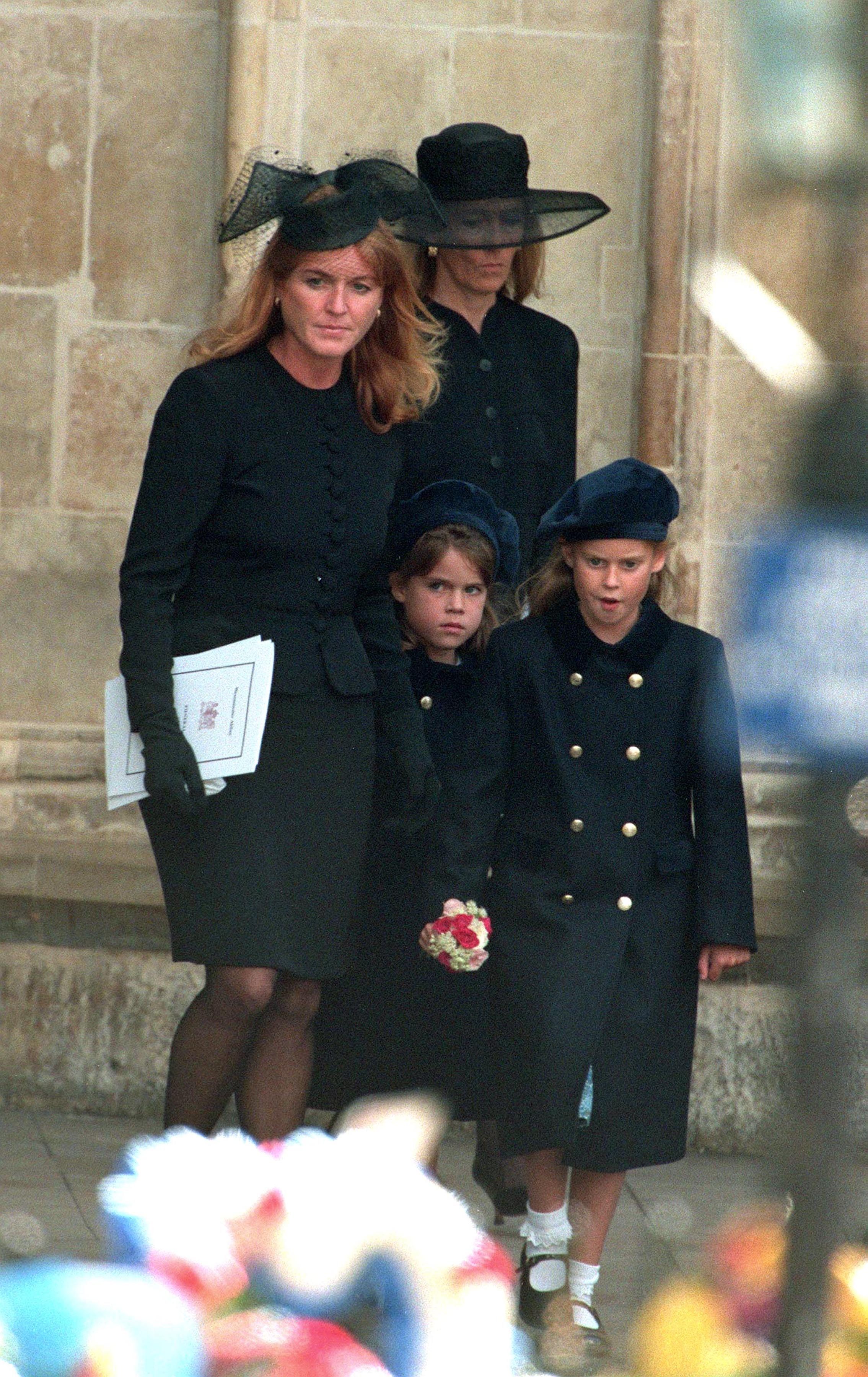 Women and children stand solemnly at a formal event, wearing dark clothing and hats, holding papers and flowers against a stone wall backdrop.
