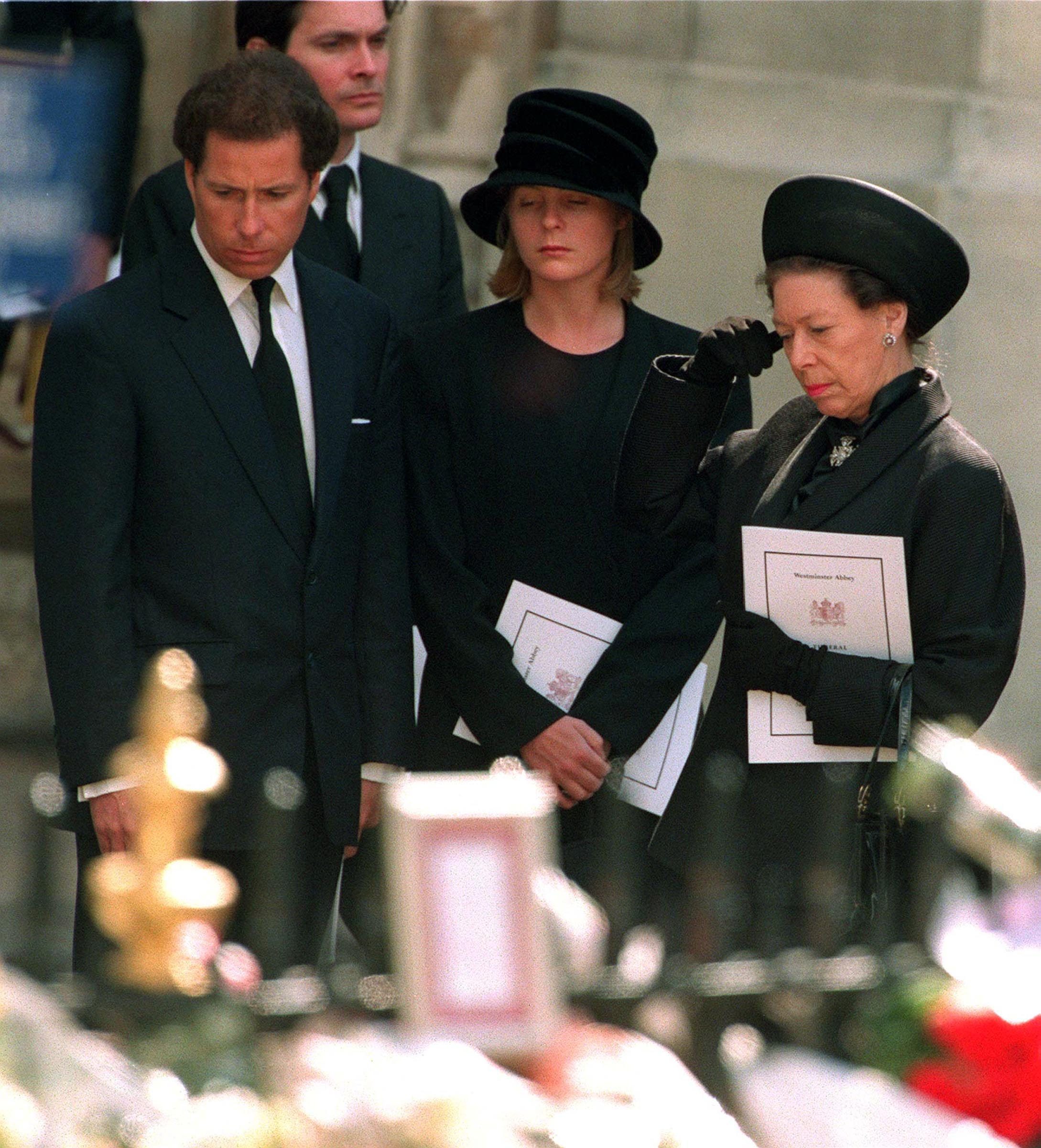 Three people stand solemnly, holding programs at a funeral, surrounded by blurred floral tributes.