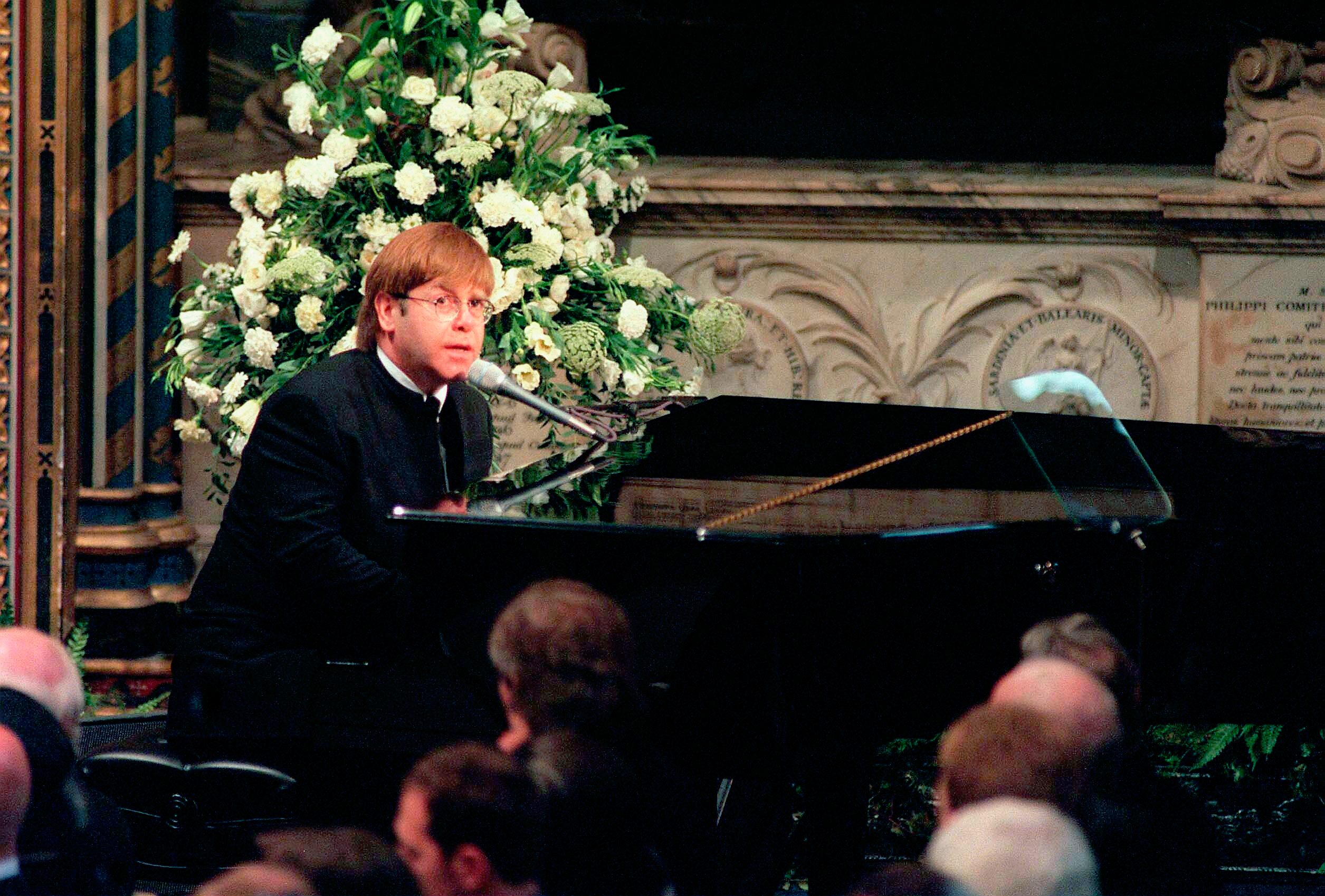 A person plays piano and sings, surrounded by ornate woodwork and floral arrangements, with an audience of people in formal attire watching.