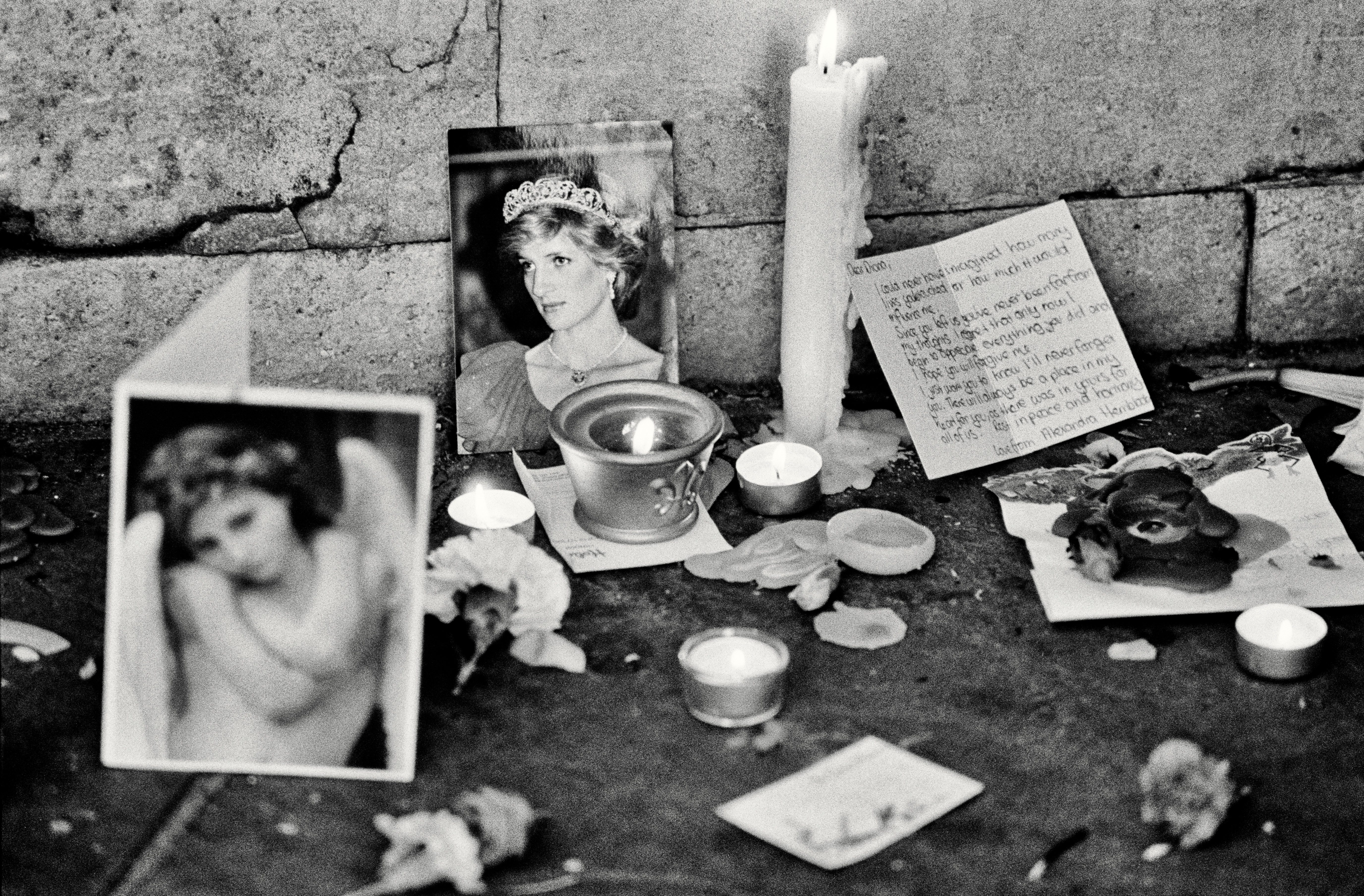 Candles flicker beside photographs and handwritten notes, creating a memorial on the ground against a stone wall.