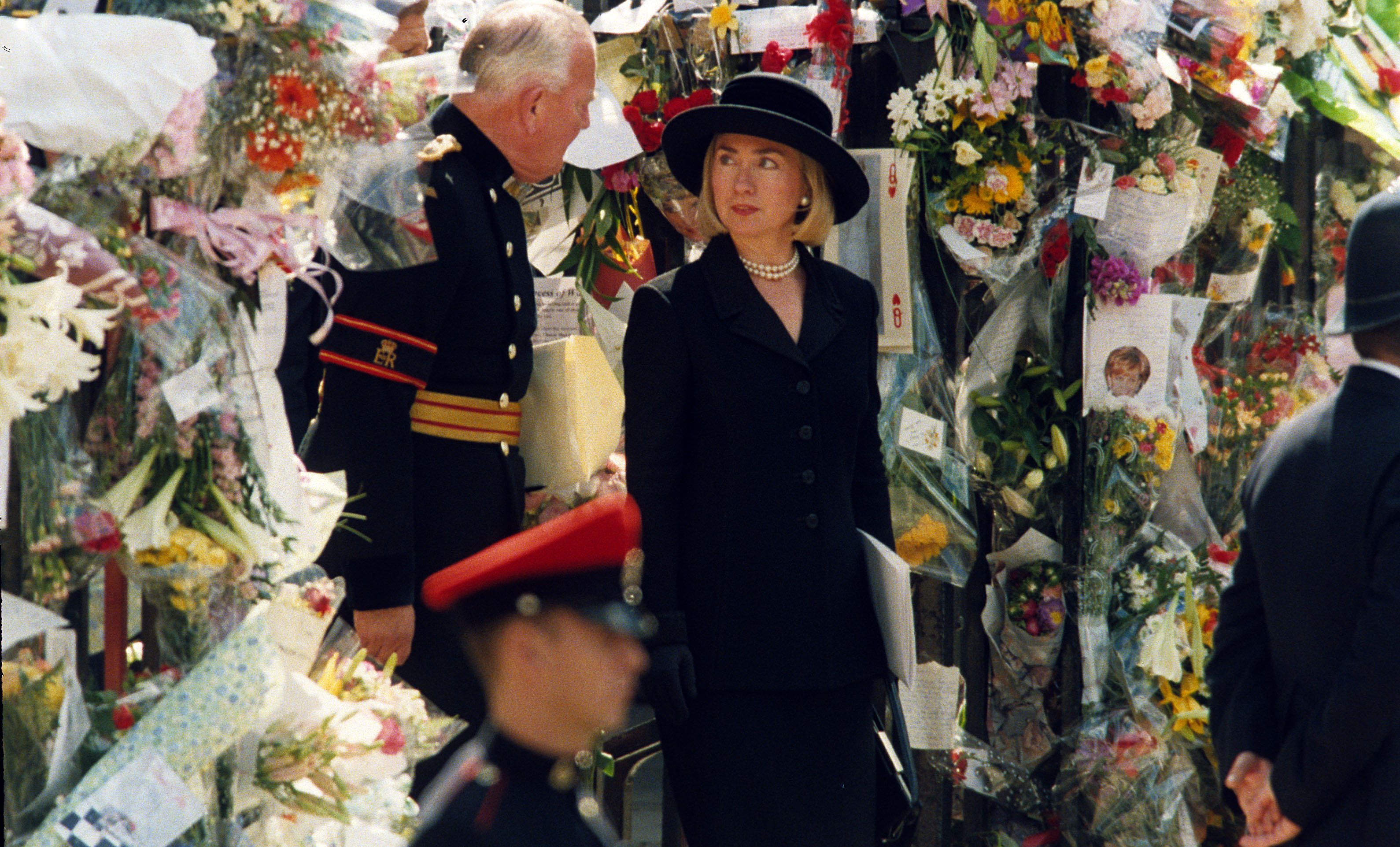 Two people in formal attire converse amid a backdrop of numerous floral tributes, creating a somber, respectful atmosphere.