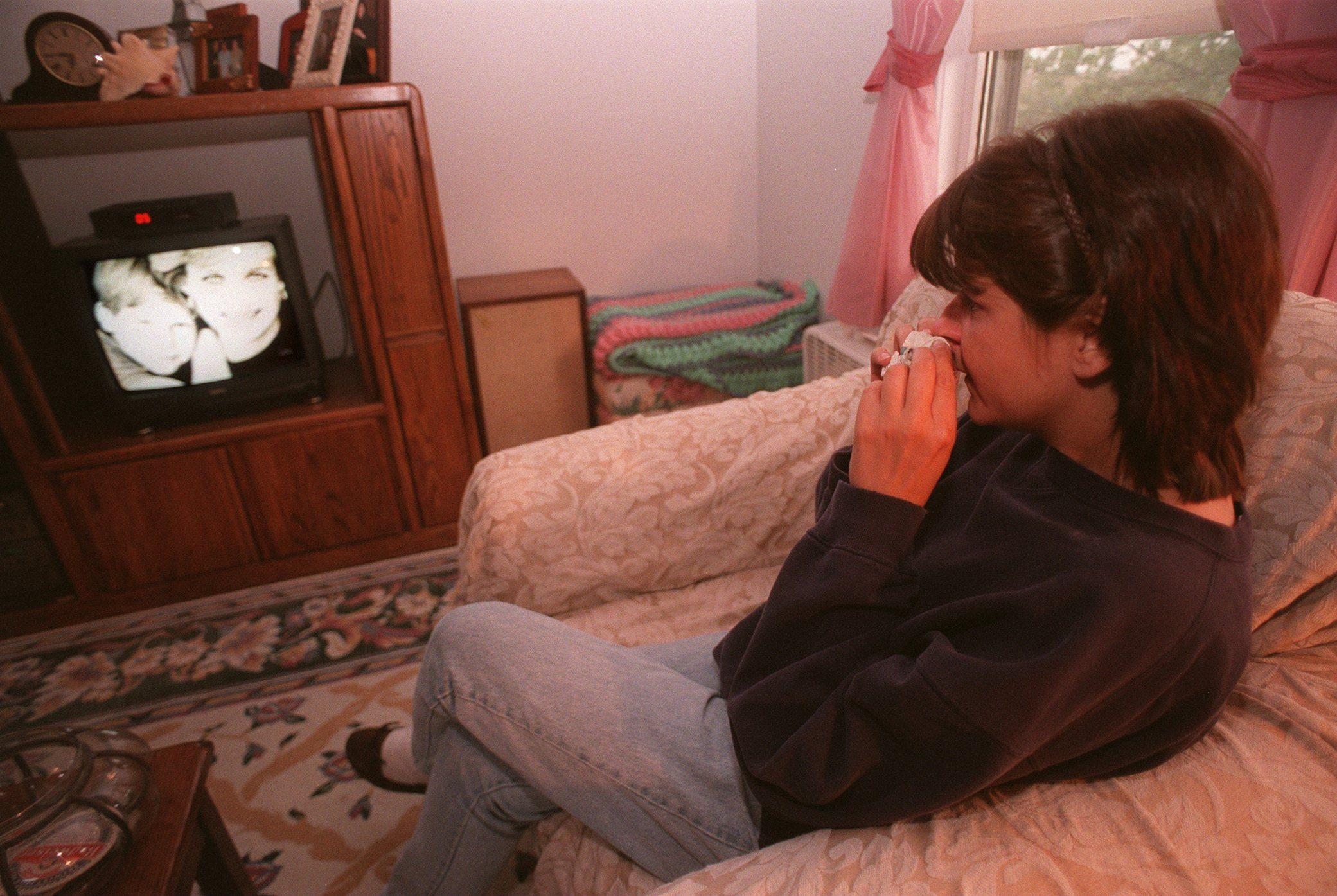 Woman sits crying into tissue, watching TV; living room features floral rug and pink curtains.