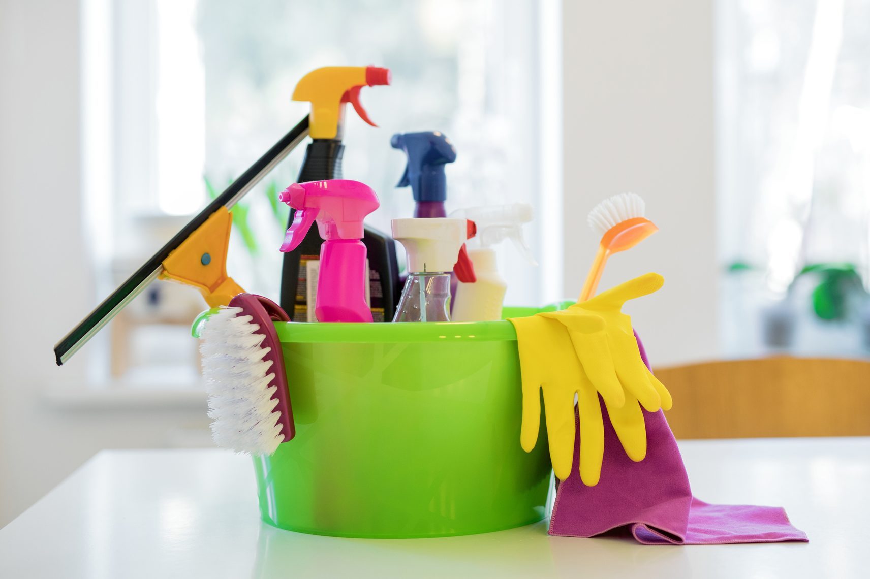 Green bucket holds cleaning supplies, including spray bottles, brushes, gloves, and cloth, on a white table in a bright room.