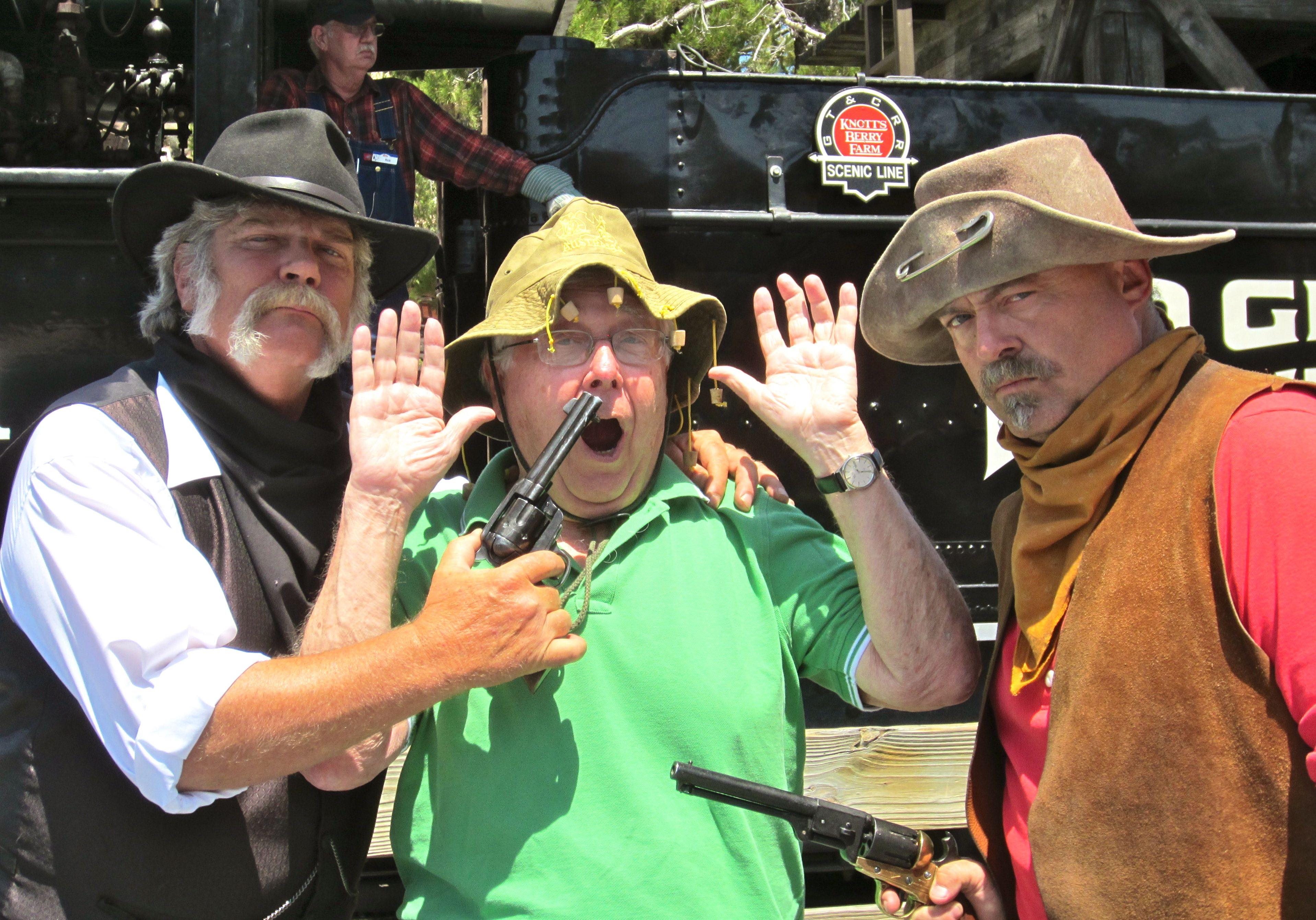 Man raises hands, appearing surprised as two others point old-fashioned guns at him. All are dressed in Western attire, standing near a train.