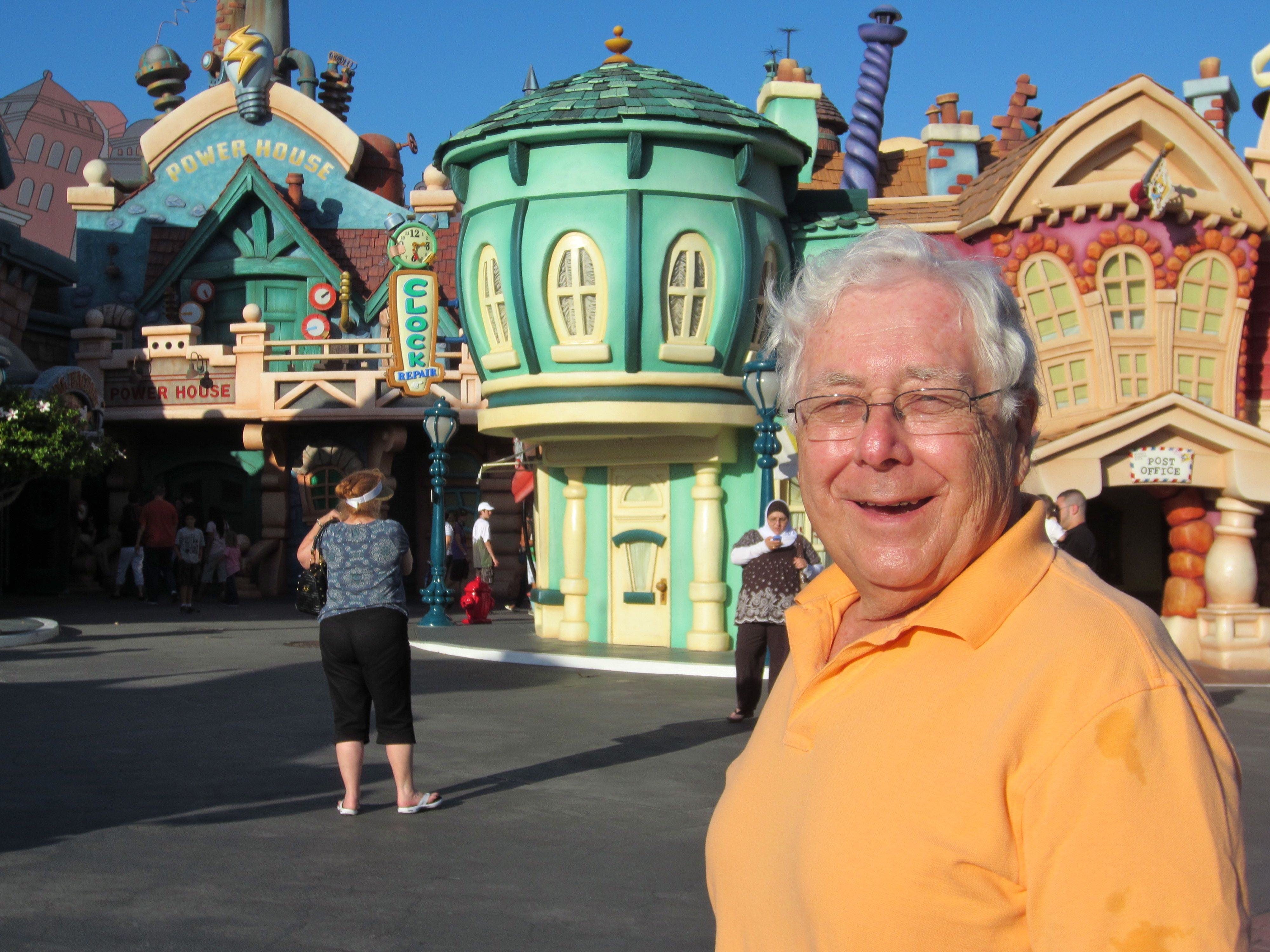 Smiling man stands; colorful, whimsical buildings in the background; people walk nearby; signs read "Power House" and "Clock Repair." Bright, sunny day.