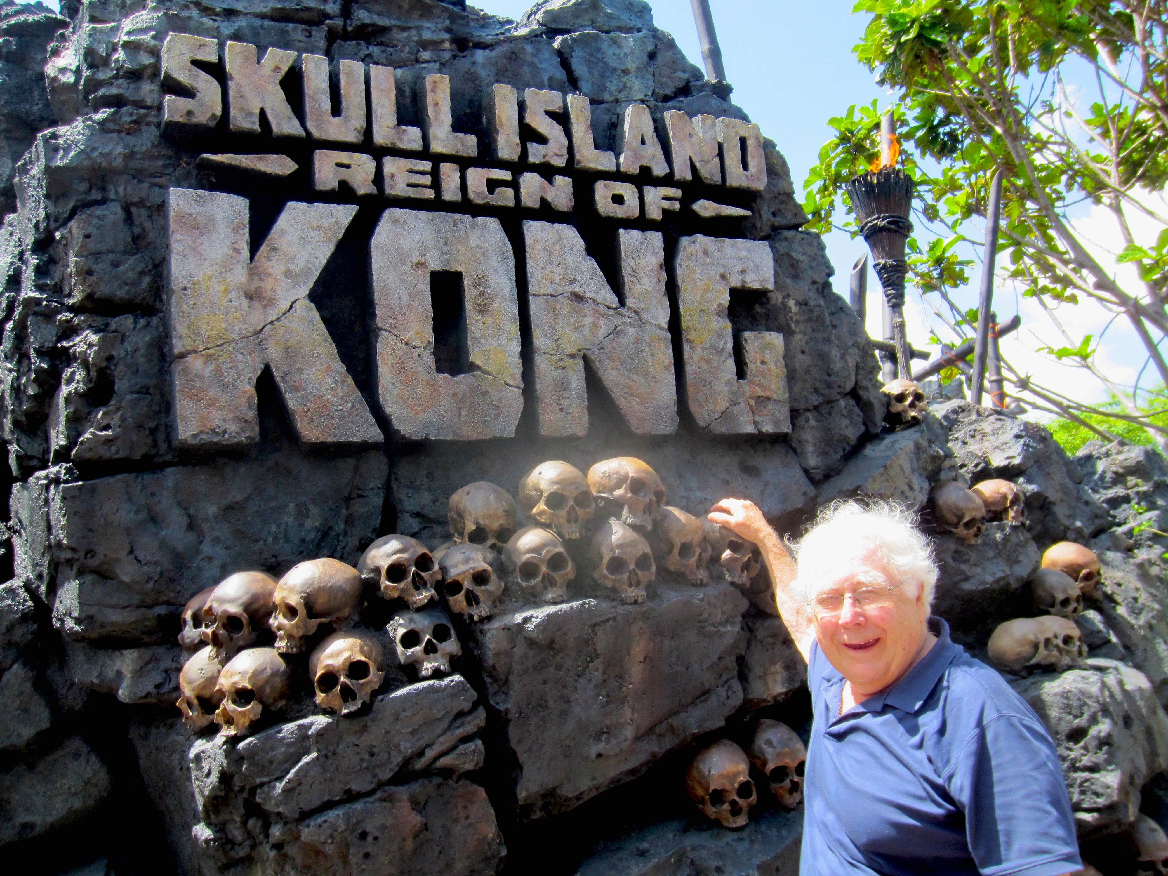 A man poses beside a stone wall with skulls and a sign reading "Skull Island Reign of Kong" amidst tropical foliage and torches.