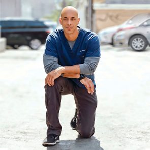 A person kneels on a sunlit street, wearing scrubs and sneakers, with parked cars blurred in the background.