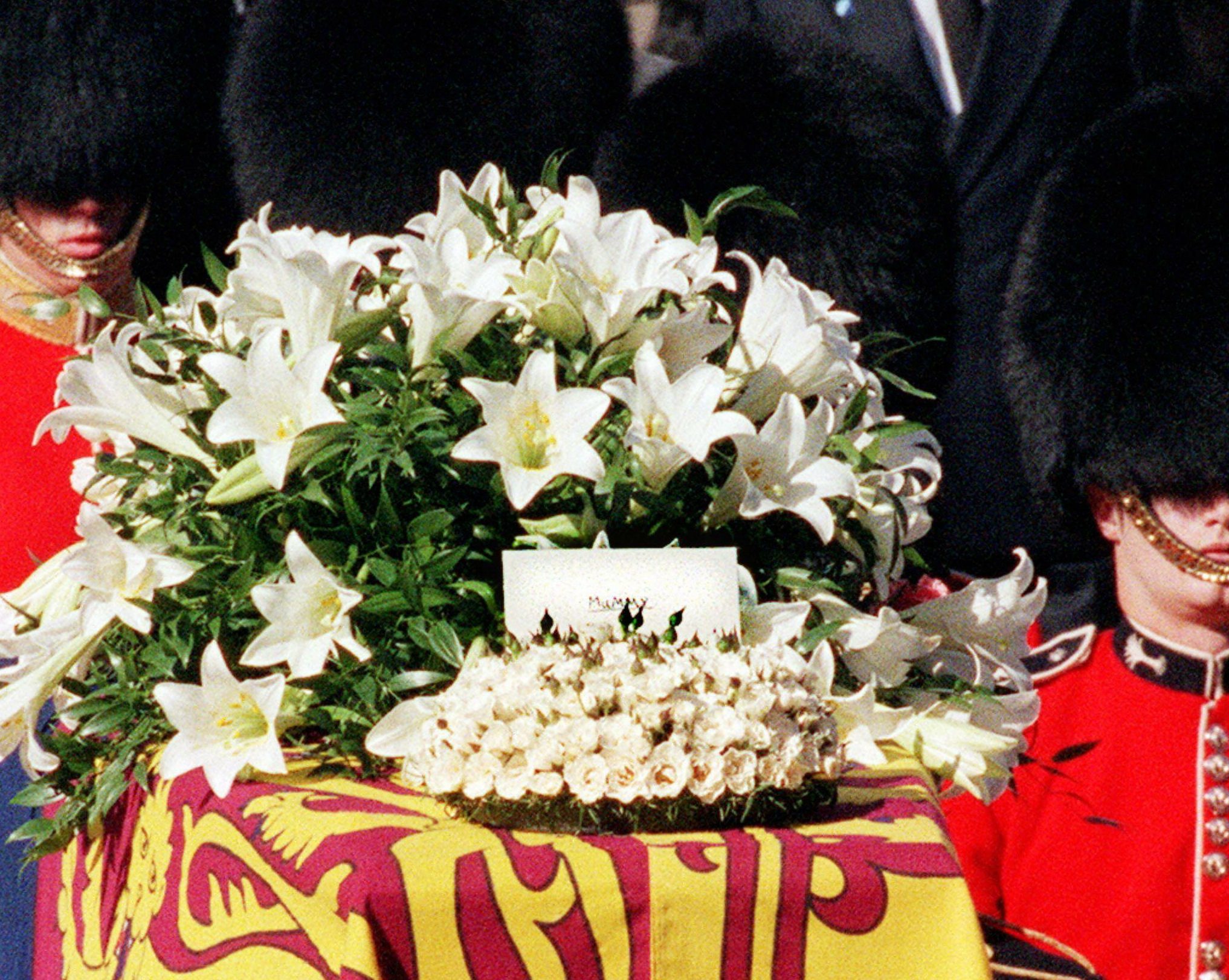 White lilies adorn a draped casket surrounded by guards in red uniforms. A card with "Mummy" is visible among flowers.