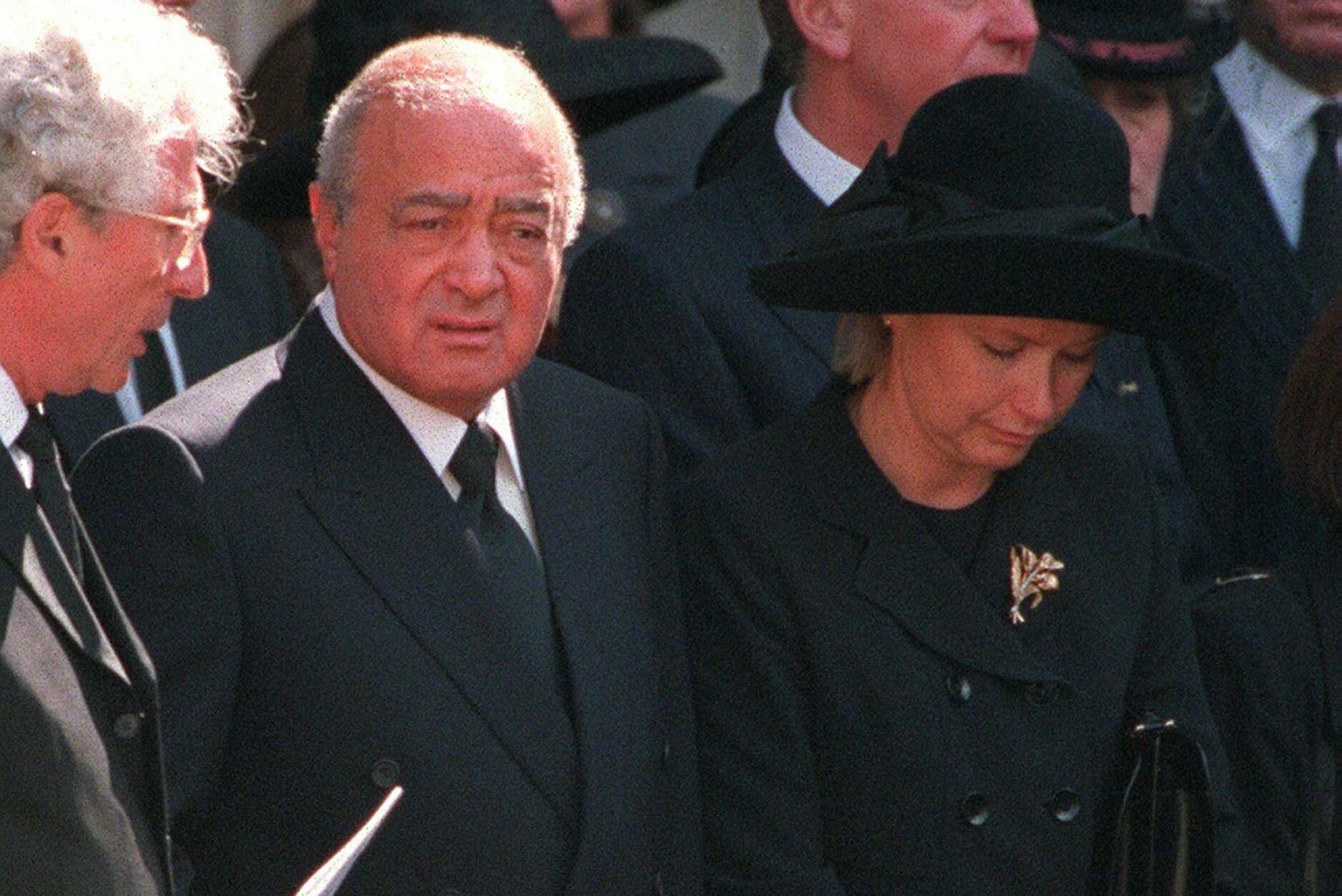 A man in a suit stands solemnly among people in formal black attire, holding papers, in an outdoor setting.