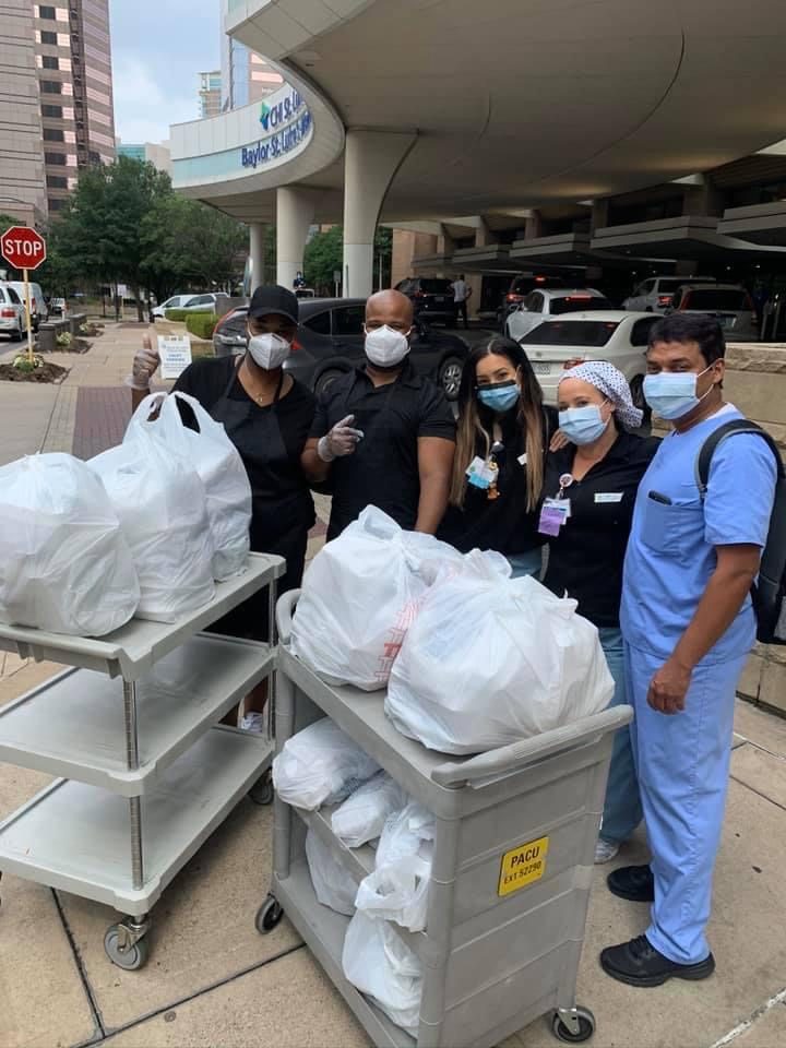 People in masks stand by carts filled with bagged items outside a hospital entrance, showing support. Signs and trees are visible in the background.