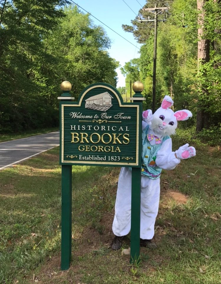 A person in a bunny costume waves beside a "Welcome to Our Town, Historical Brooks Georgia" sign, amidst a wooded roadside area.