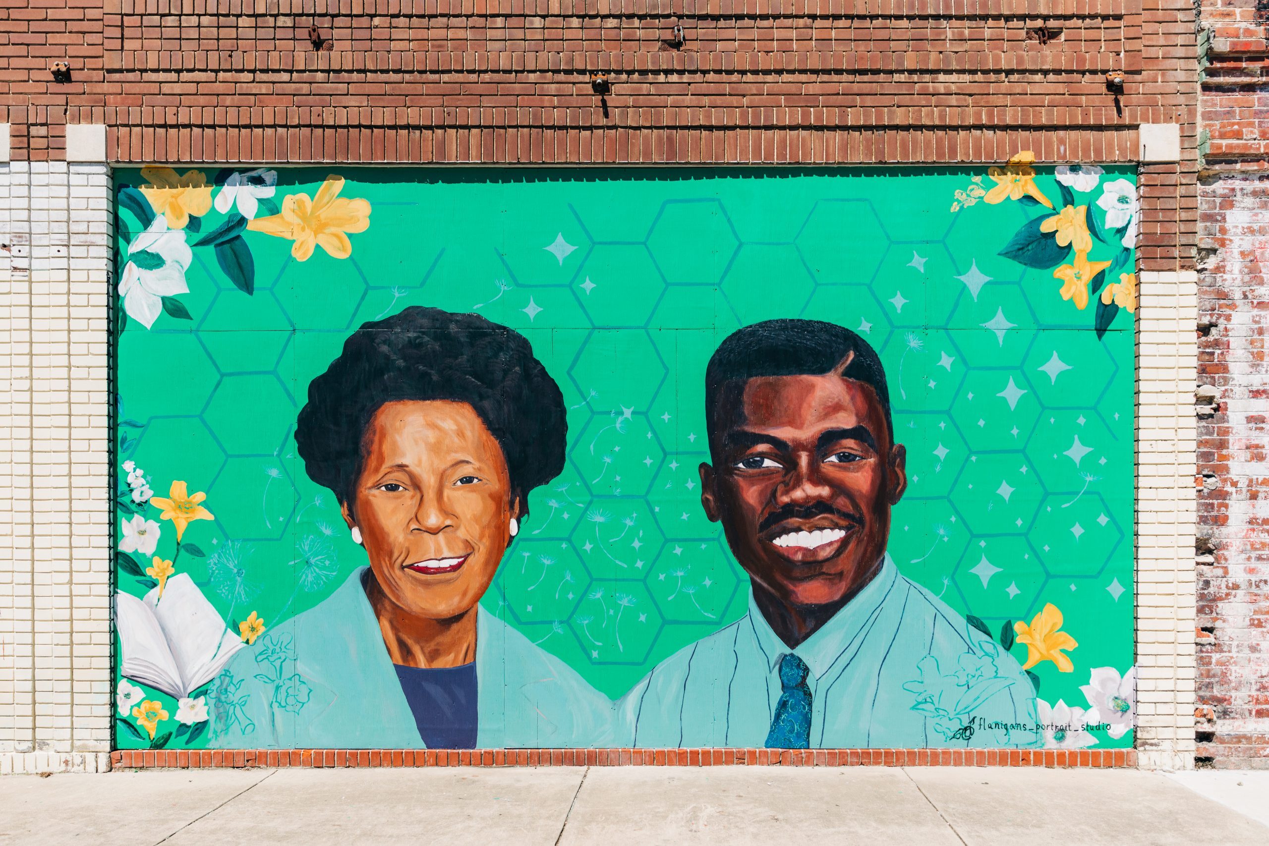 Mural depicts two smiling portraits on a green, floral background with hexagonal patterns on a brick wall.