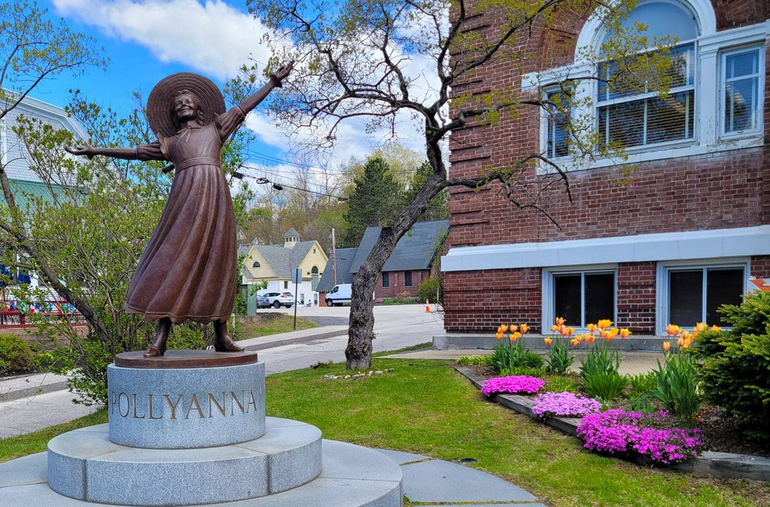 Bronze statue of a girl joyfully poses with outstretched arms on a round base. Surrounded by brick buildings and gardens, text reads "POLLYANNA."