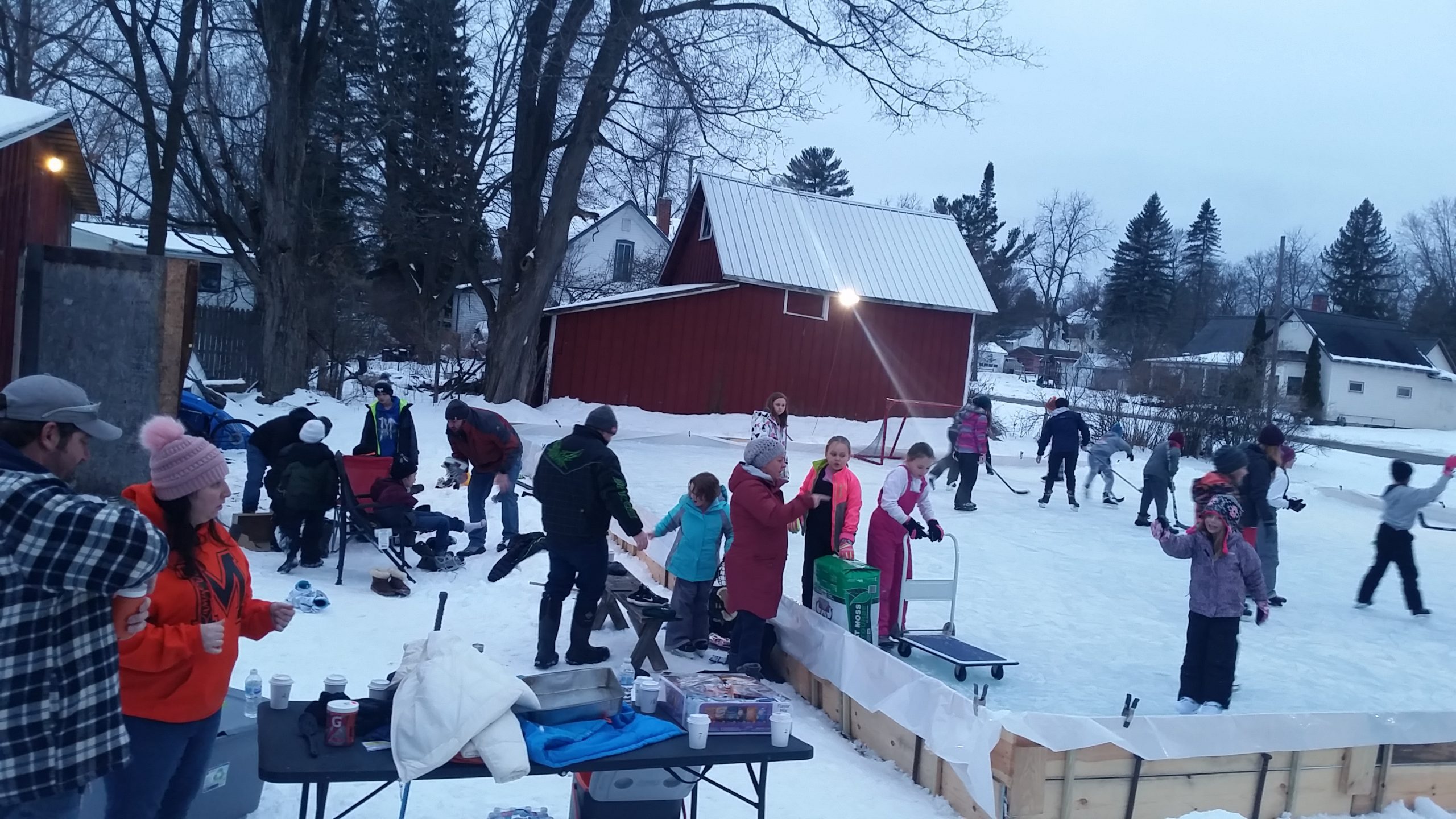 People gather on a snowy ice rink; children play hockey while adults converse by a table with hot drinks. A red barn and trees surround.