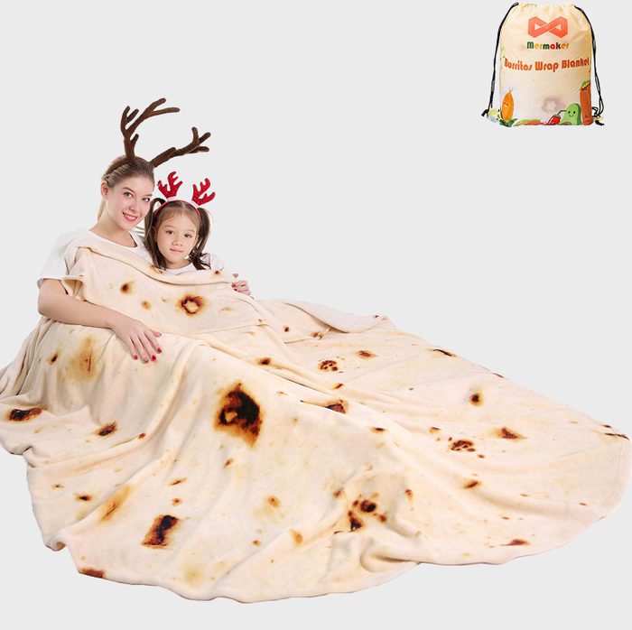 Two people sit under a large tortilla-patterned blanket, wearing reindeer antlers, smiling in a studio setting. A drawstring bag is labeled 