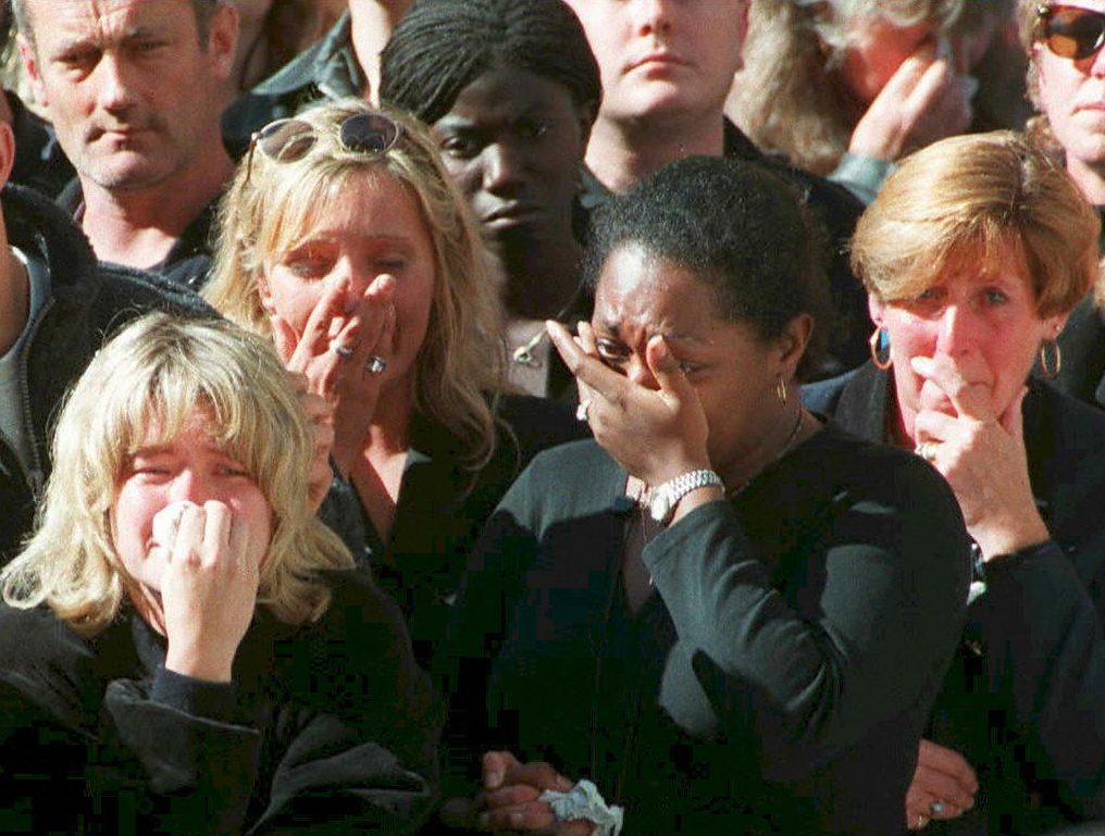 People are crying and covering their faces; they appear distressed in a crowd, wearing dark clothing, likely at a somber or emotional event.