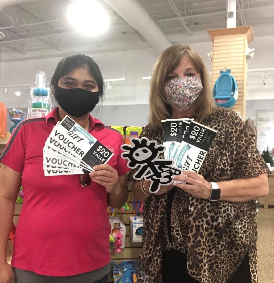 Two people holding gift vouchers in a store, standing side by side, wearing masks amidst colorful merchandise.