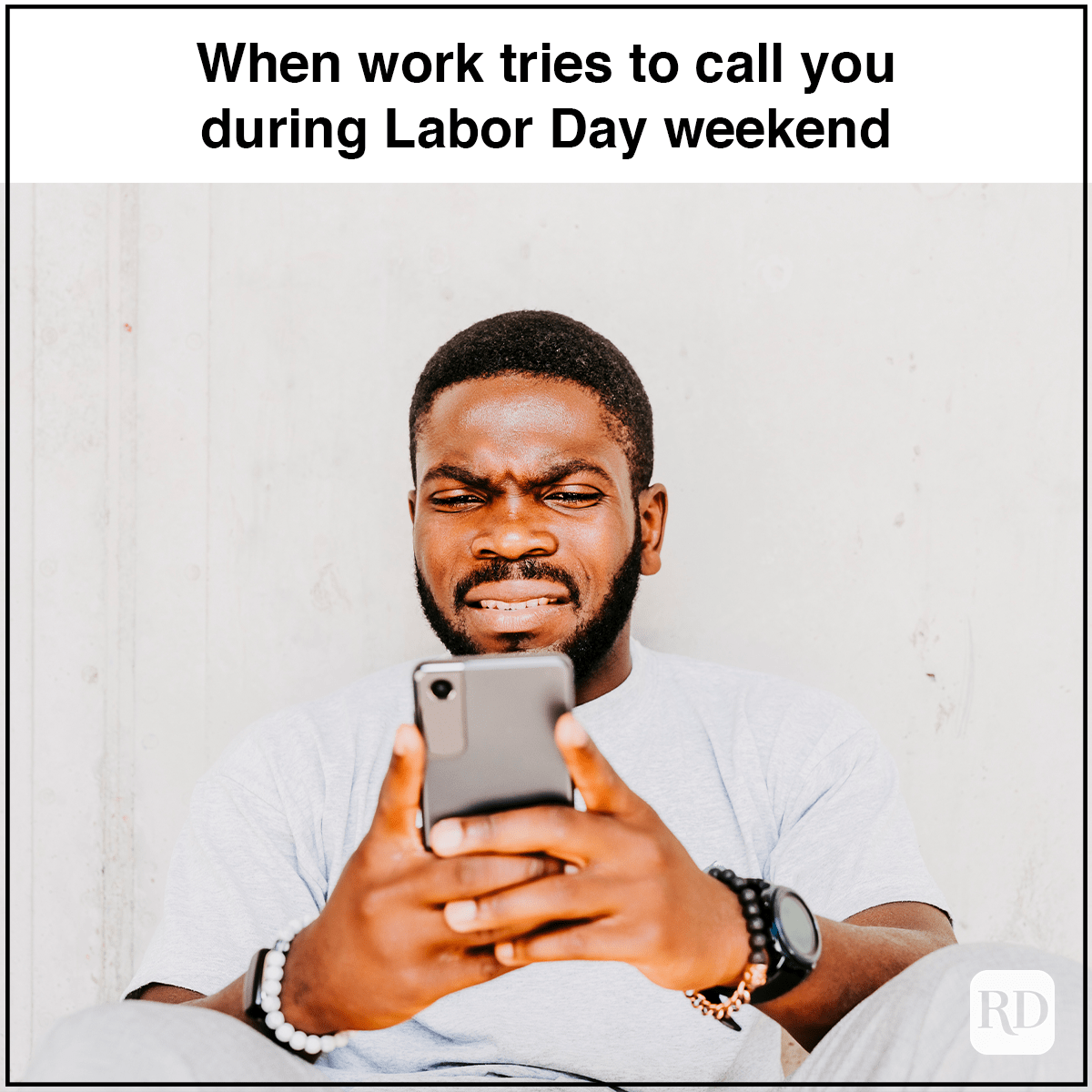 Man grimaces while looking at phone, sitting against a light wall. Text reads: "When work tries to call you during Labor Day weekend."