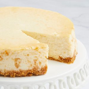 Cheesecake on a white stand, missing a slice, with a creamy top and crumbly crust, set against a light marble background.