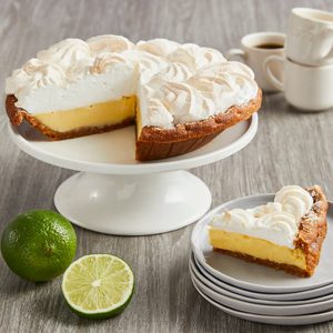 Pie sits on a stand with a slice removed; surrounded by limes and stacked plates, in a dining setting with coffee cups.