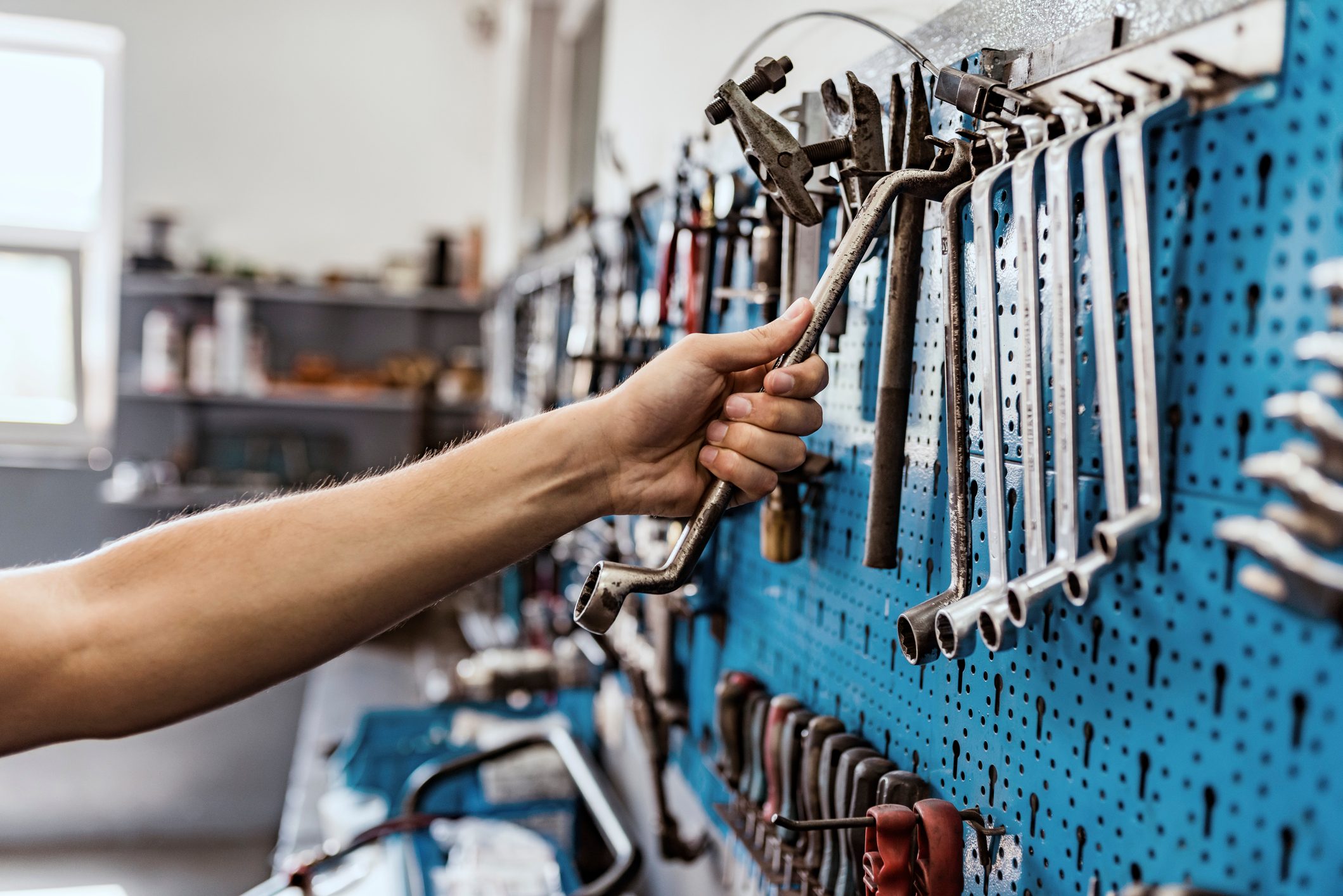A hand selects a metal tool from a blue pegboard, surrounded by various hanging tools in a workshop setting.