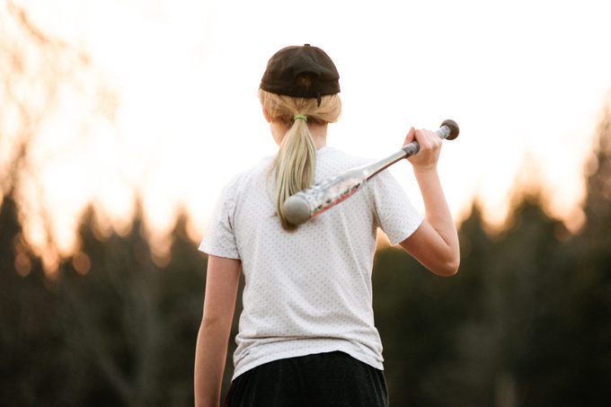 Girl with baseball bat over her shoulder in rural field, rear view