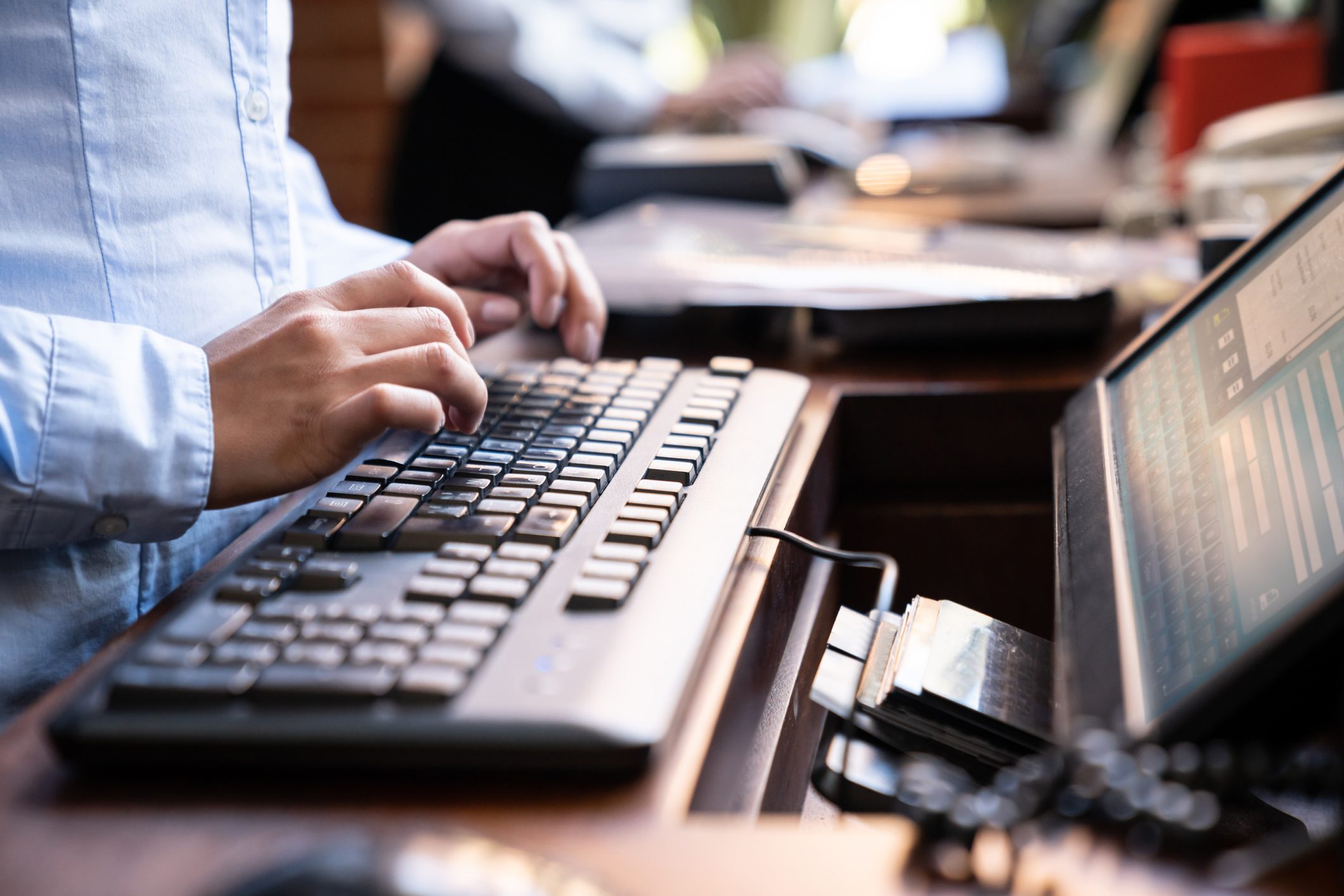 Hands typing on a keyboard in an office setting, with a monitor showing information and blurred background.