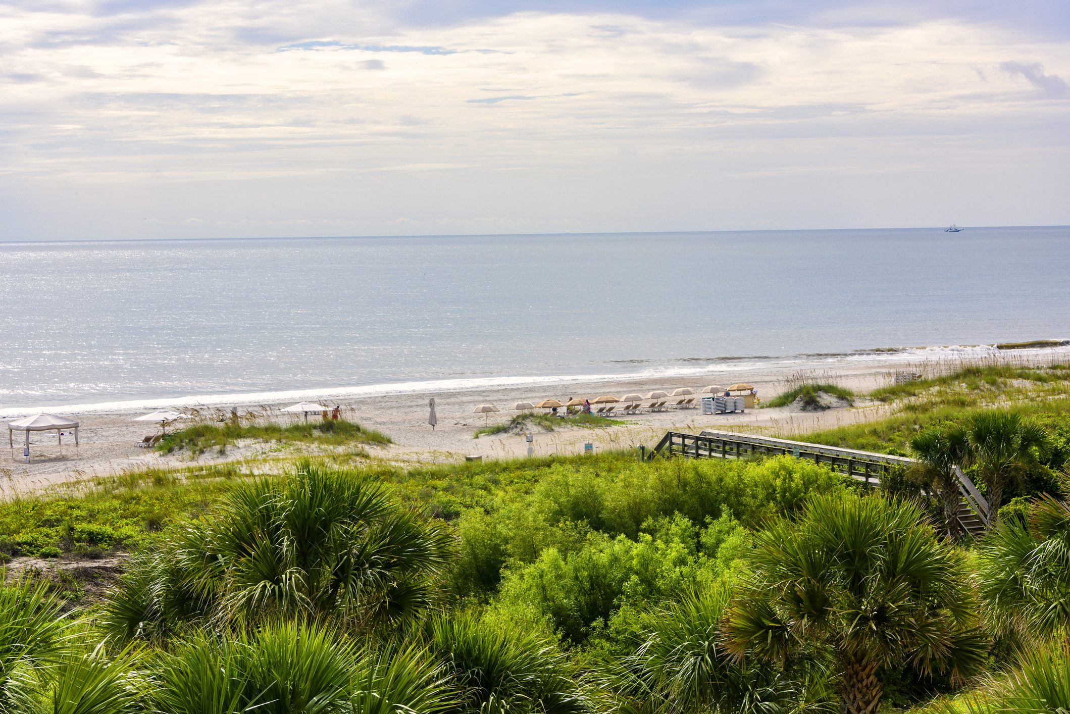 Beach chairs line the sandy shoreline under a cloudy sky, with lush greenery and a wooden pathway in the foreground.