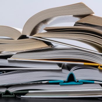 Books lie open, pages fanned outward, stacked disorderly in a well-lit room.