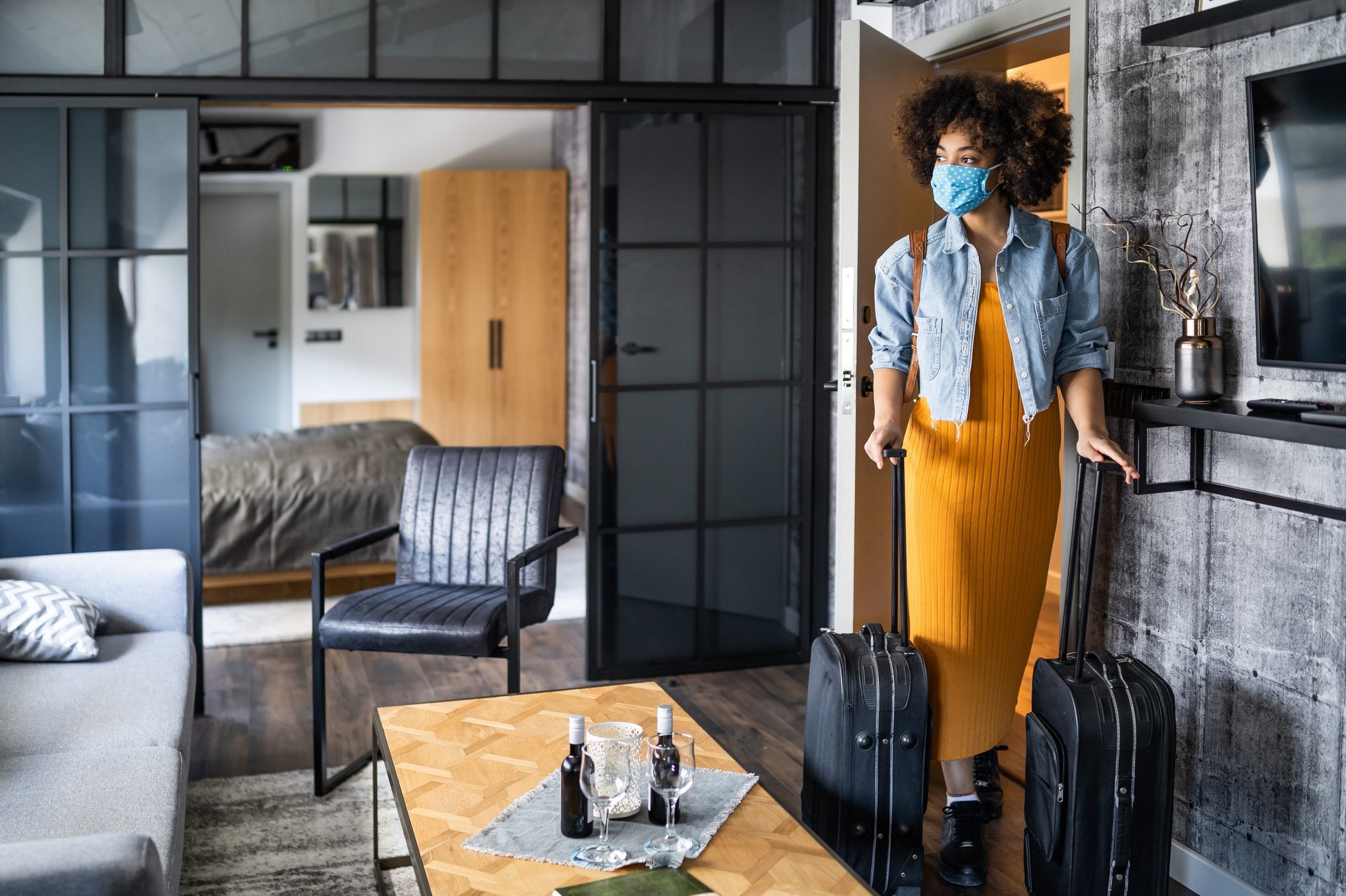 Person enters room pulling suitcases, wearing a mask and denim jacket, next to a modern living area with a table and black chair.