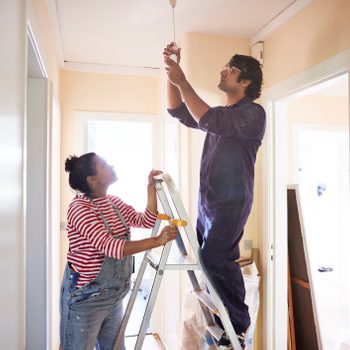 A person on a ladder installs a light bulb while another assists, inside a brightly lit room with light-colored walls.