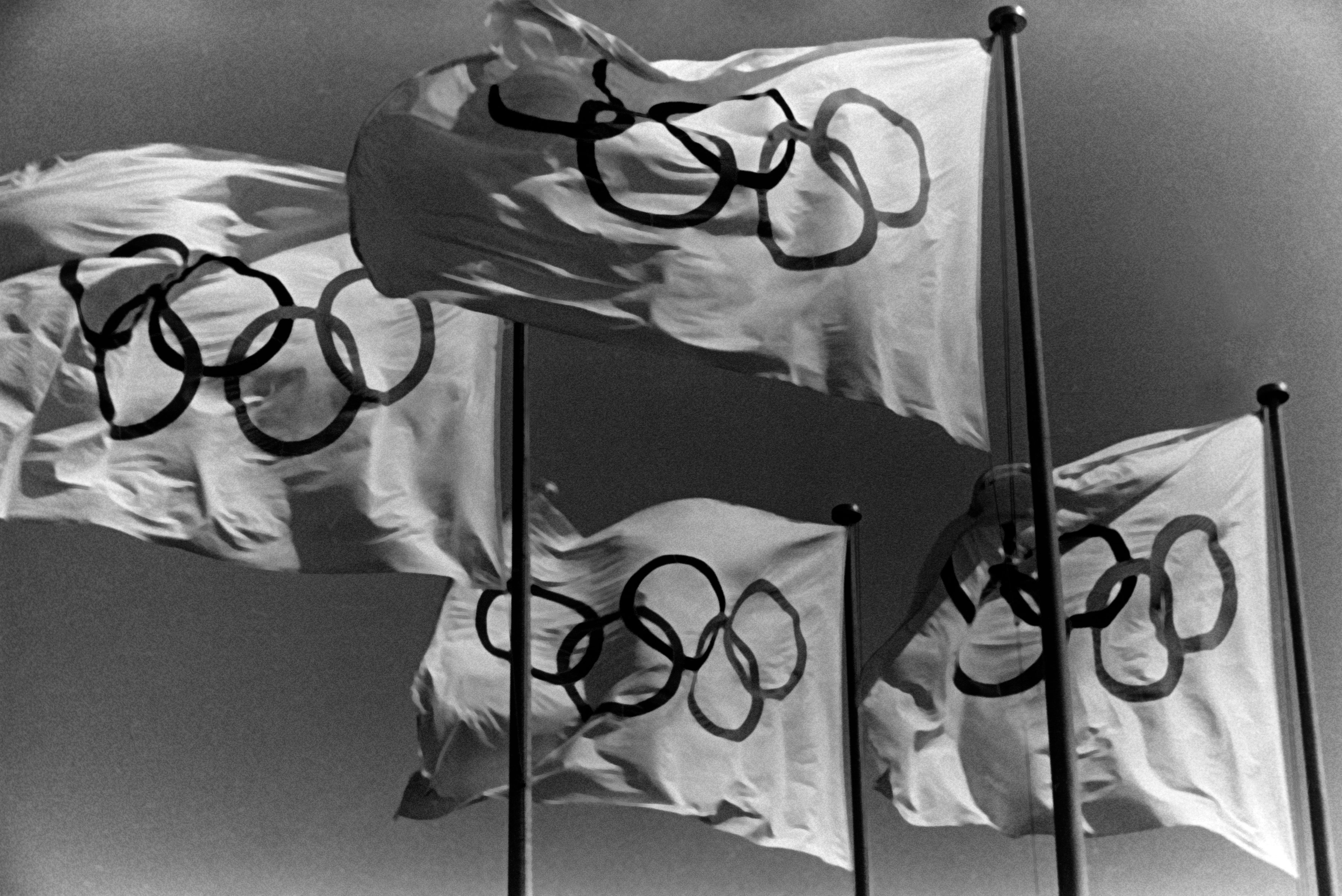 Flags with Olympic rings wave in the wind against a clear sky.