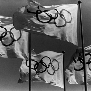 Flags with Olympic rings wave in the wind against a clear sky.