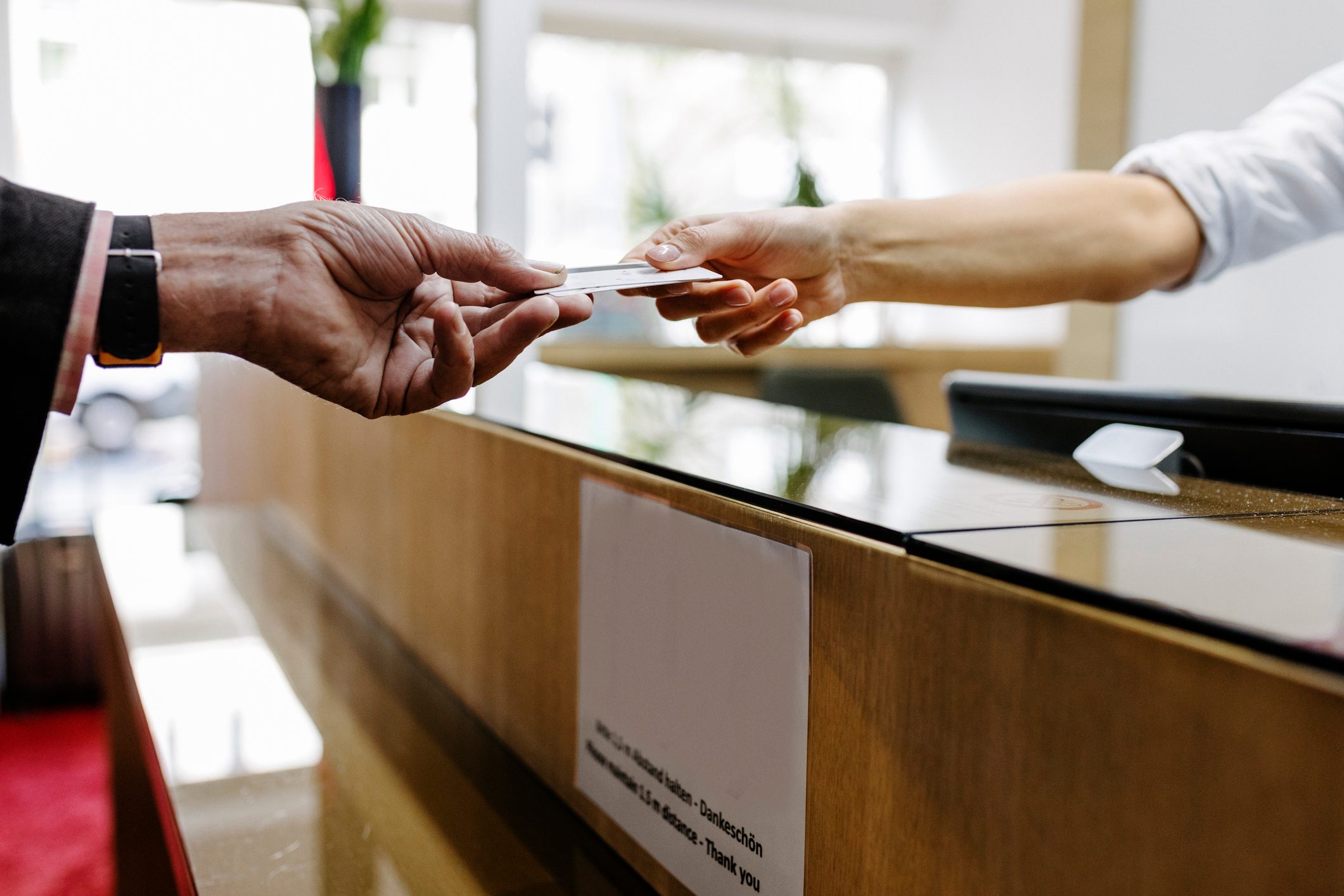 A hand, holding a card, reaches across a reception desk towards another hand in a modern, bright interior. Sign on desk: "13.5 cm."