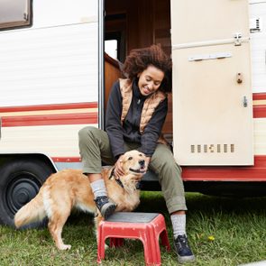 Person sitting, petting a happy dog outside an RV. A red step stool rests on grass beside them.