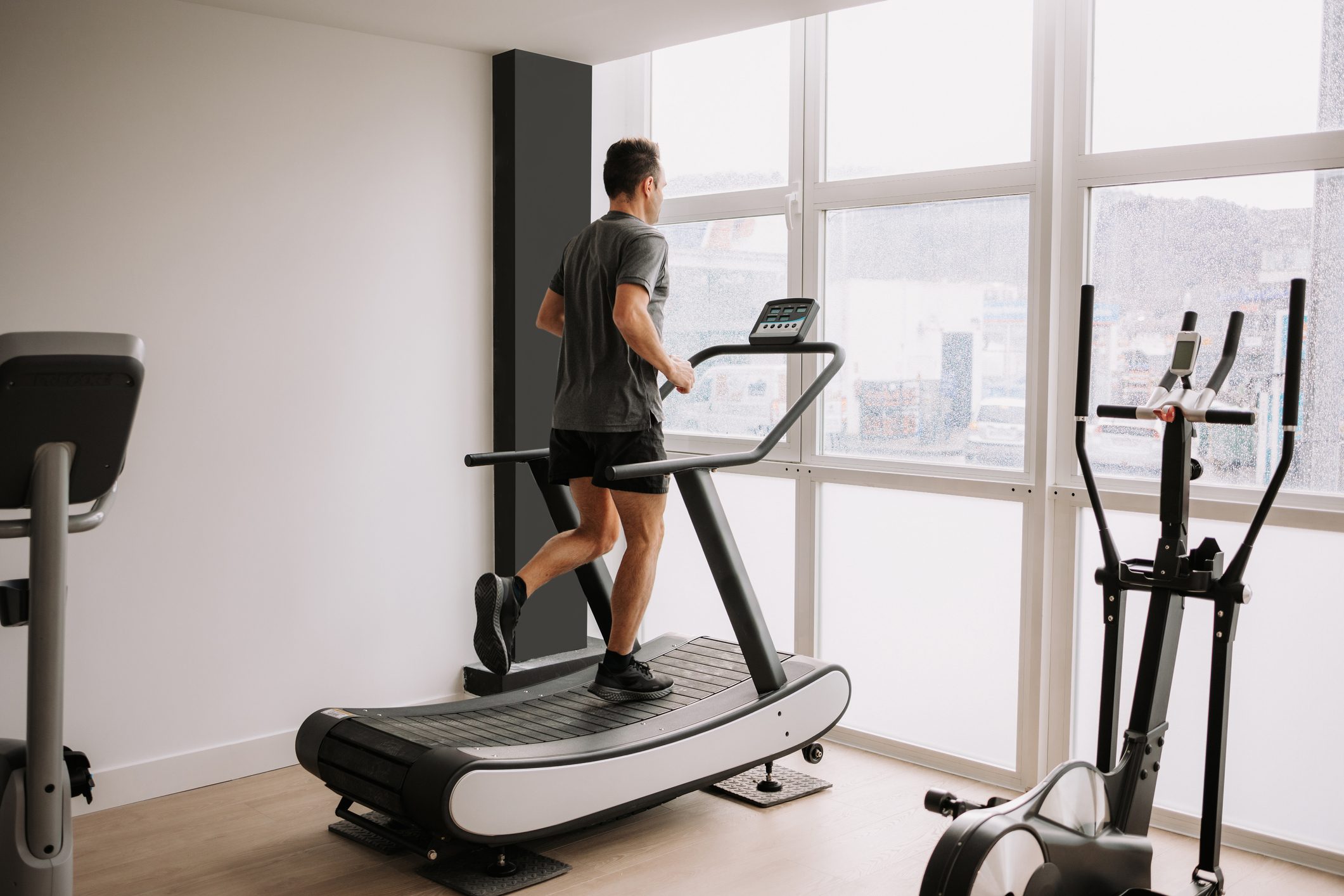 Person running on a treadmill facing large windows in a bright gym, with exercise equipment nearby.