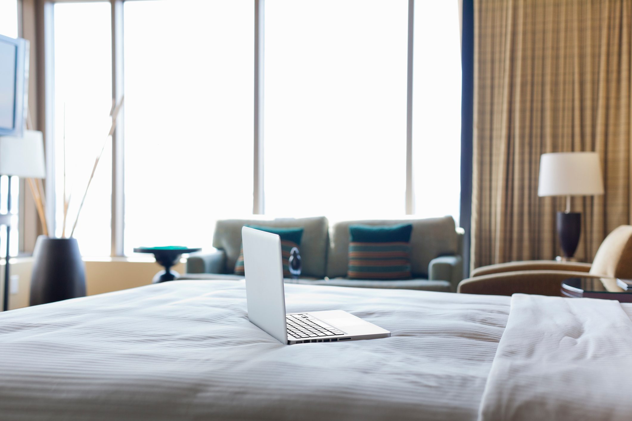 Laptop sits open on a bed in a bright, modern room with large windows and soft furniture.