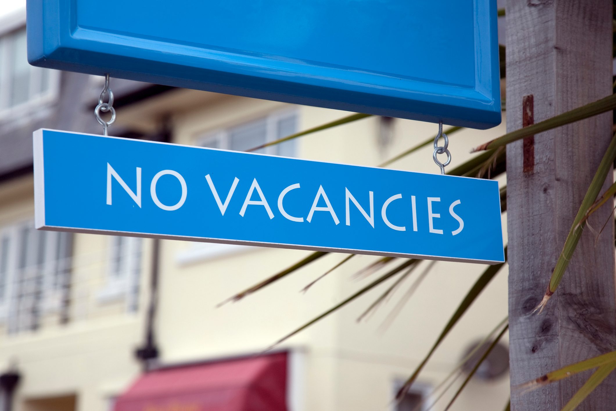 A blue sign reads "NO VACANCIES," hanging outside beside a wooden post, with blurred buildings in the background.