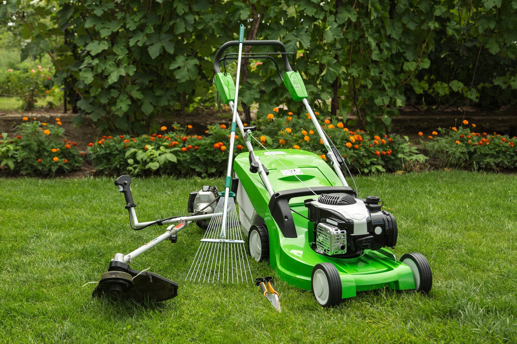 Lawnmower, rake, and trimmer rest on grass amidst flower beds and lush greenery in a garden setting.