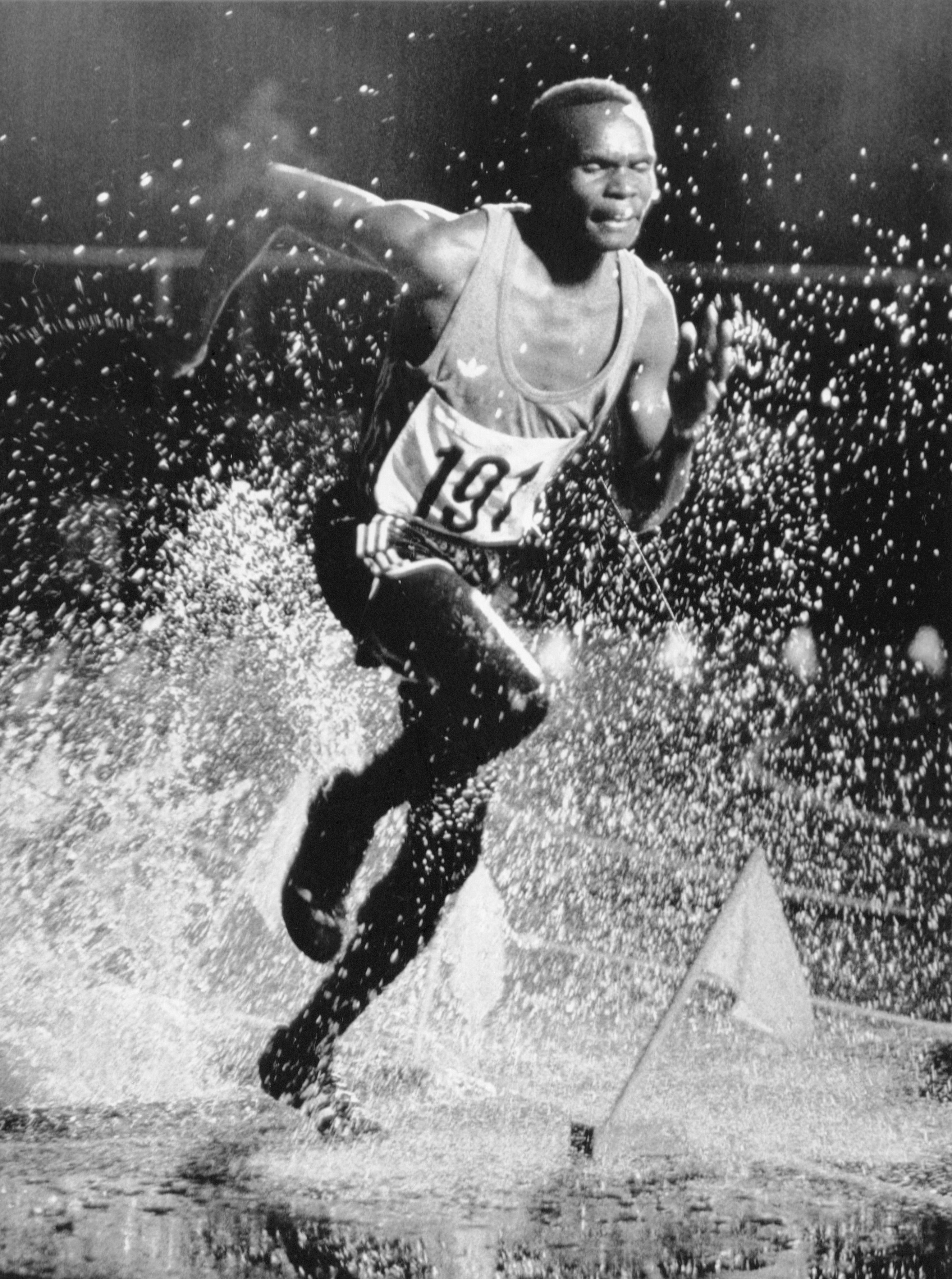 Runner strides through water, splashing dramatically, wearing athletic gear with number 191; captured in black-and-white, suggesting action and intensity in a wet race environment.