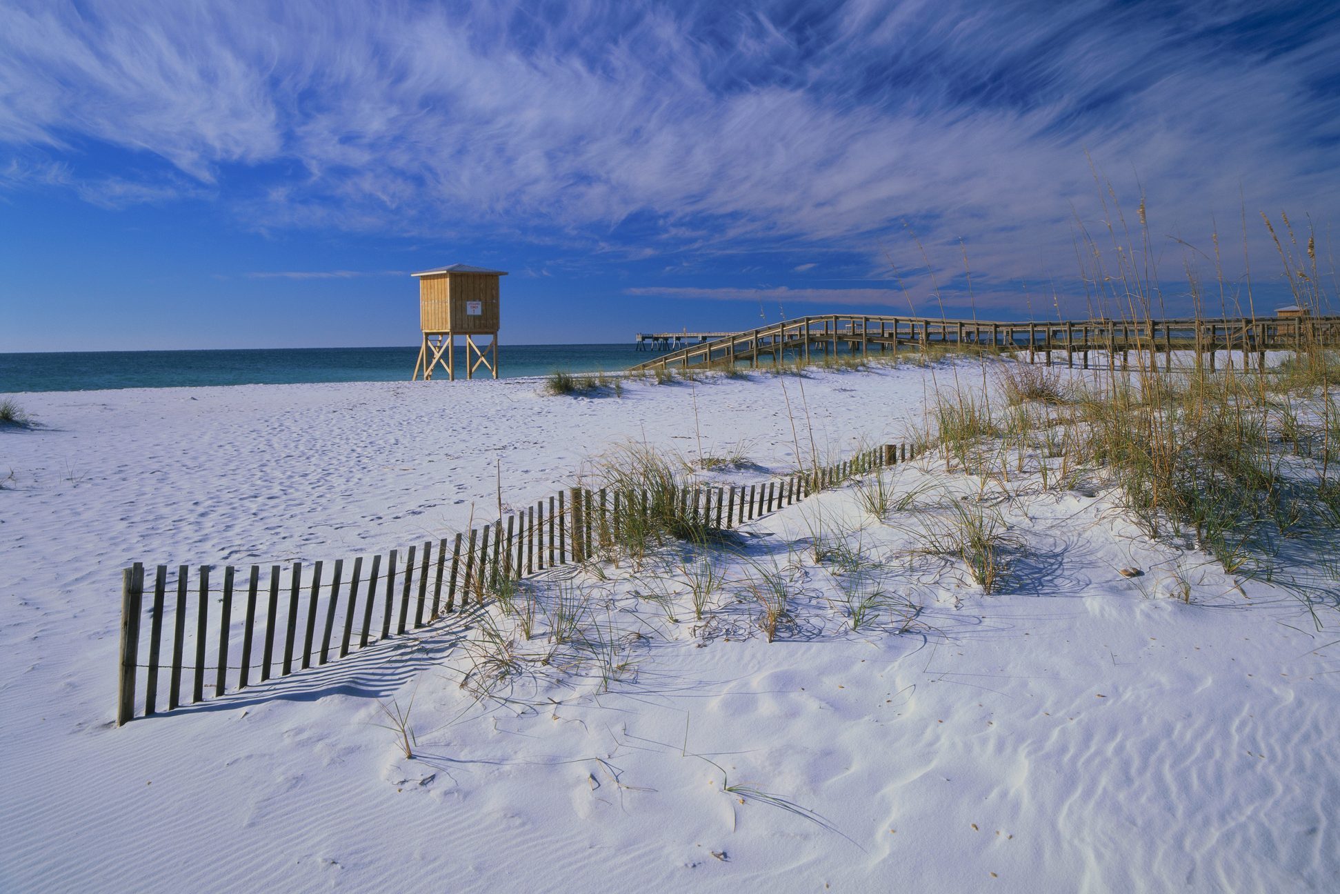 Wooden lifeguard tower stands on white sandy beach, near a fenced dune, under a clear, blue sky with wispy clouds.