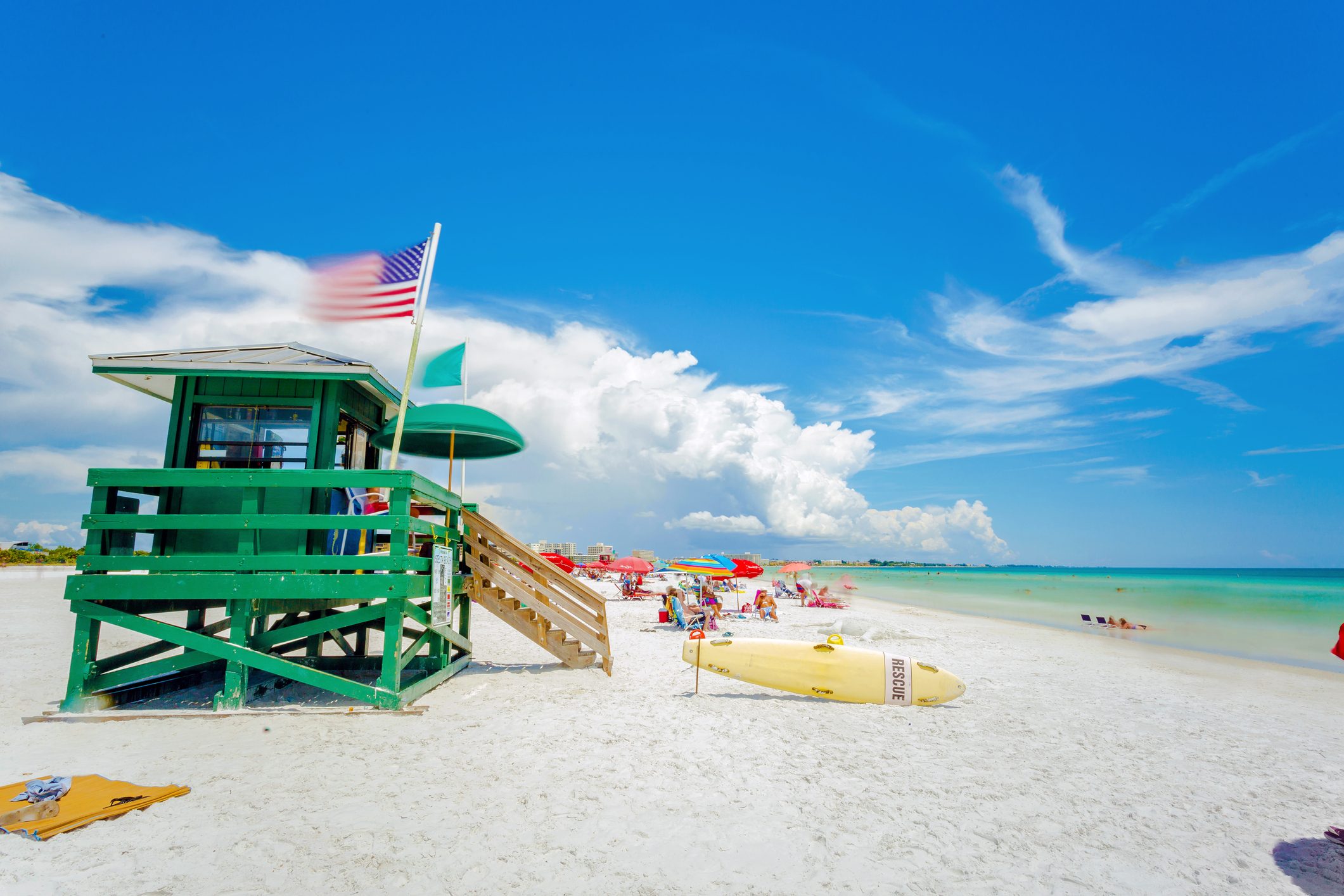 Lifeguard hut stands with flags fluttering; beachgoers relax under colorful umbrellas on a bright, sunny day near the ocean.