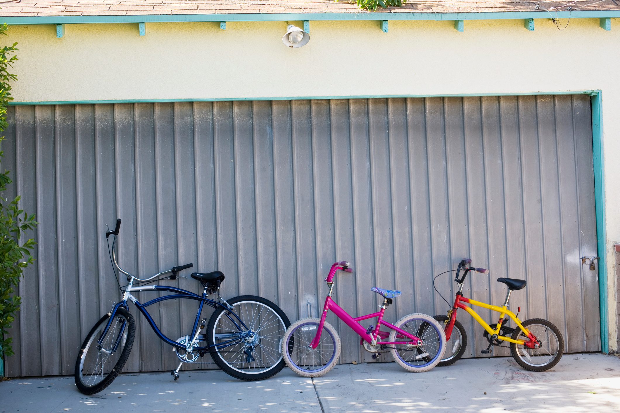 Bicycles lean against a closed garage door, with three different-colored bikes on a sunlit concrete driveway.