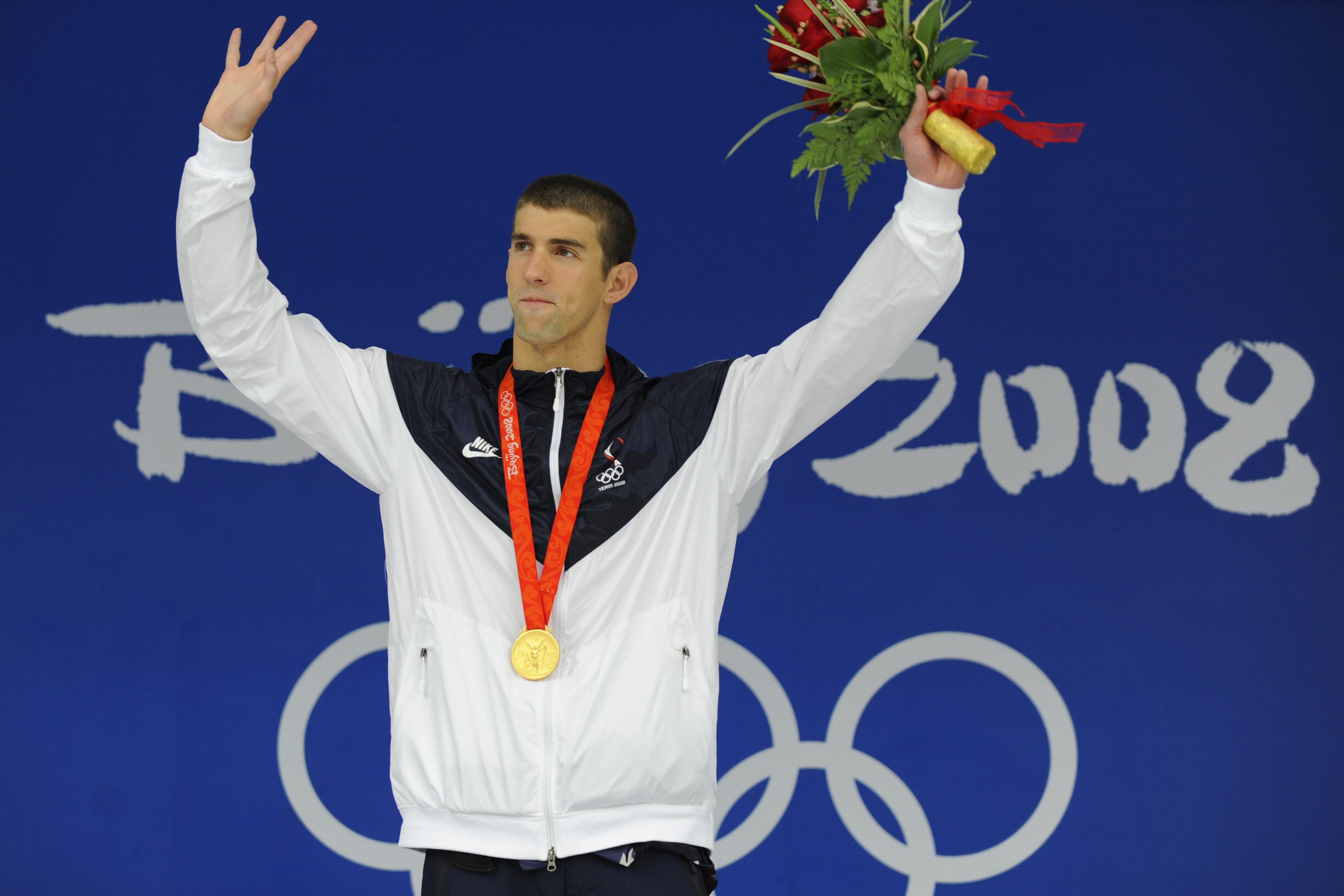 Athlete wearing a gold medal waves, holding flowers, against a blue background with "Beijing 2008" and Olympic rings.