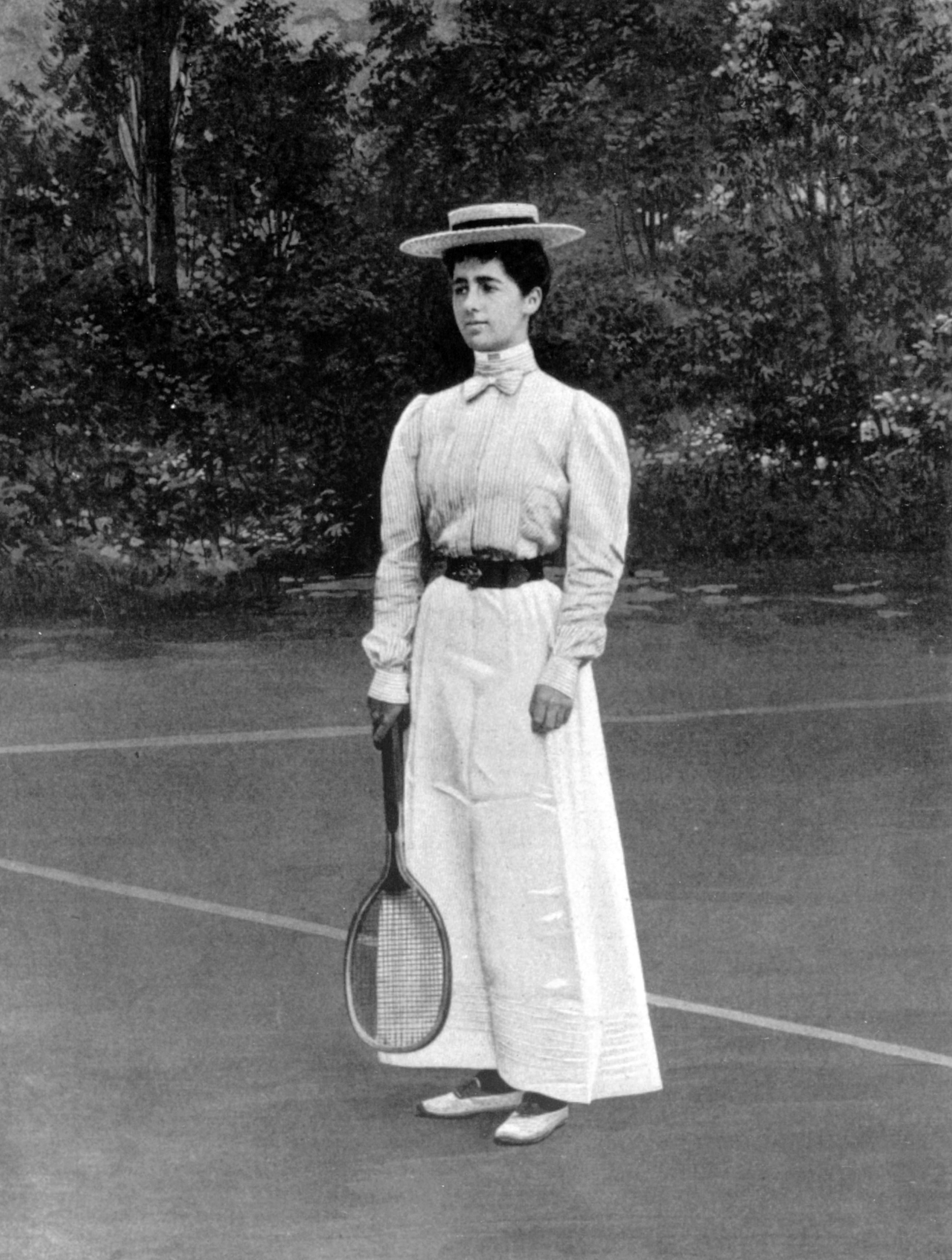 Woman holding a tennis racket stands on a court, wearing a long dress and hat, surrounded by trees in a vintage setting.